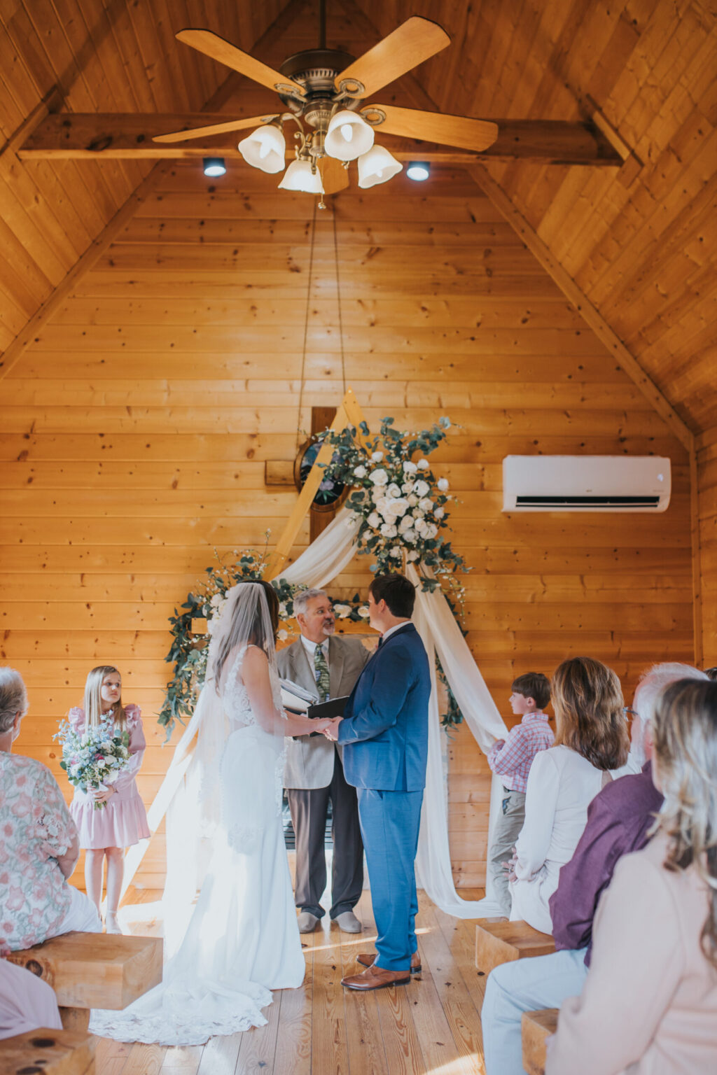 Elopement Wedding A couple, having chosen to elope, stands before an officiant in a wooden chapel, exchanging vows. The bride is in a white dress and veil, the groom in a blue suit. A young girl holding flowers and a young boy stand nearby. Guests are seated on wooden benches, with floral arrangements and draped fabric in the background. Elopements Inc