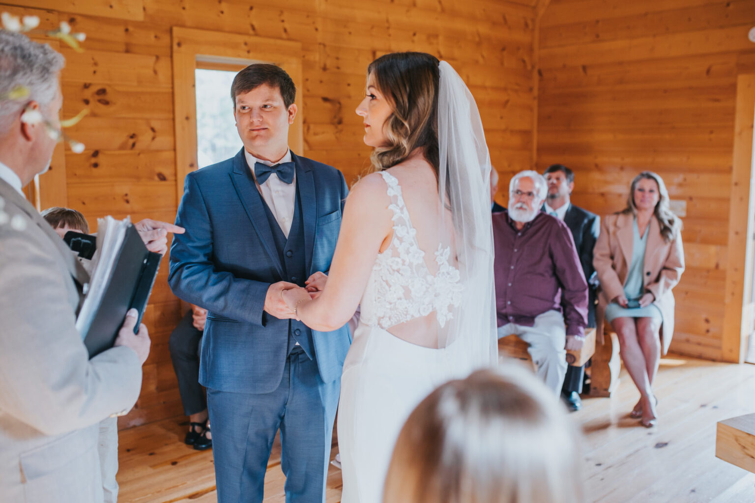 Elopement Wedding A couple is eloping in a wooden chapel. The groom, in a blue suit and bow tie, holds hands with the bride, who is wearing a white dress with lace details and a veil. A person officiating the ceremony stands to the left holding a book. Guests sit on wooden benches in the background. Elopements Inc