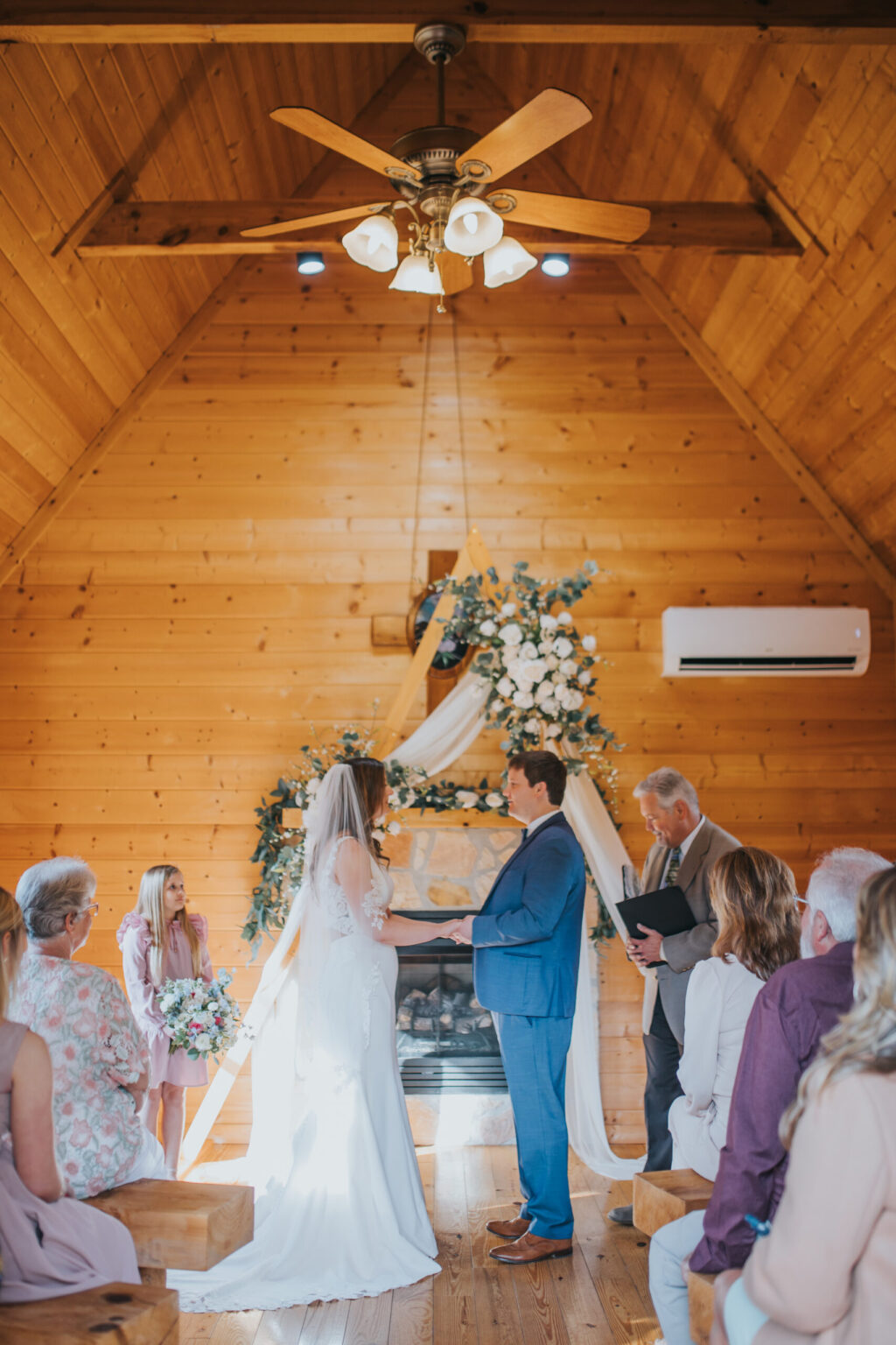 Elopement Wedding A wedding ceremony inside a wooden chapel with high, arched ceilings and a ceiling fan. The couple stands facing each other; the bride in a white gown and veil, the groom in a blue suit. An officiant stands to the right, while family and friends are seated, observing the intimate elopement adorned with floral decorations. Elopements Inc