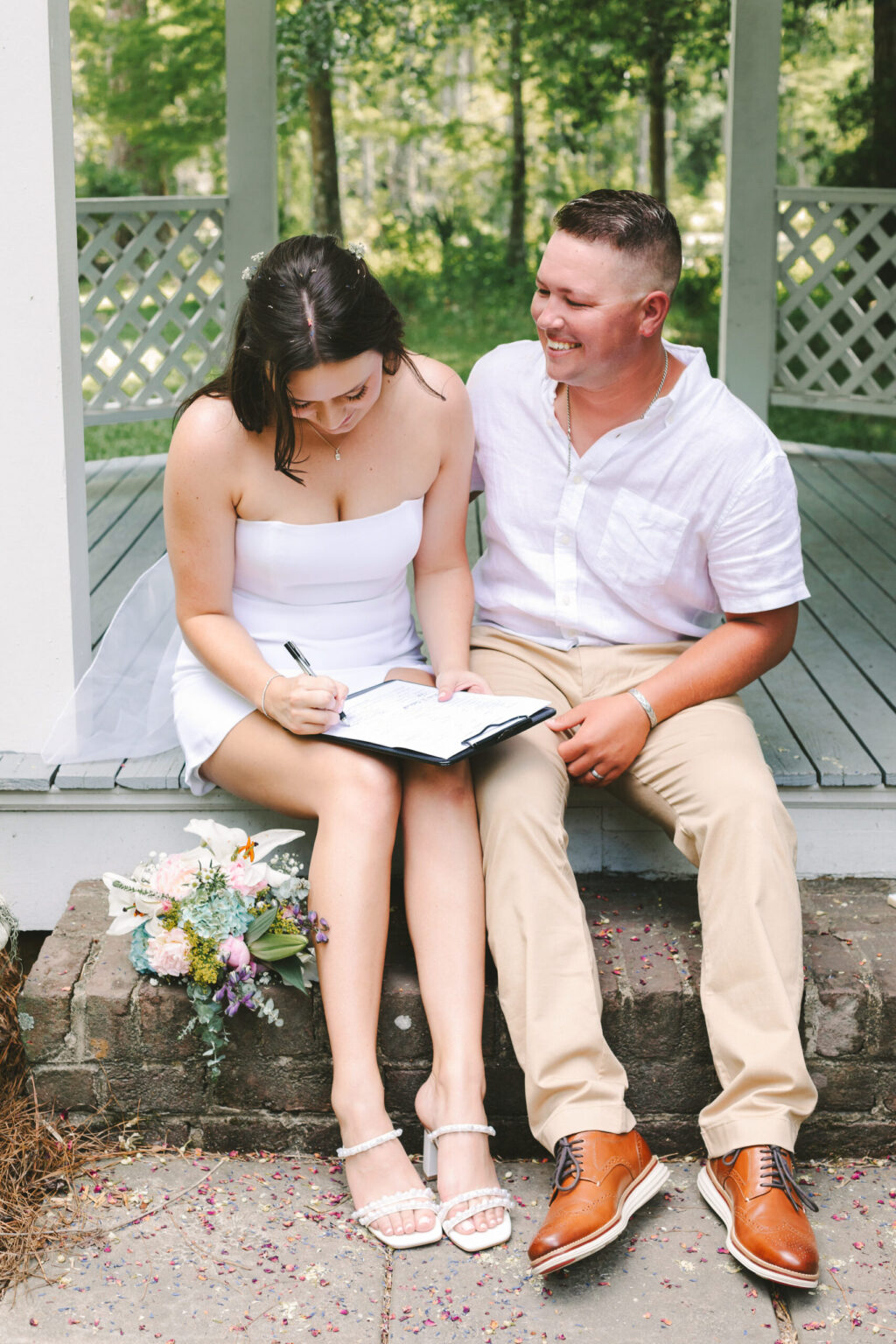 Elopement Wedding A couple elopes by sitting on a wooden platform in a gazebo. The woman, wearing a white dress and sandals, signs a document on her lap while the man, in a white shirt and beige pants, smiles at her. A bouquet of flowers rests on the ground beside them. The background is lush with green trees. Elopements Inc