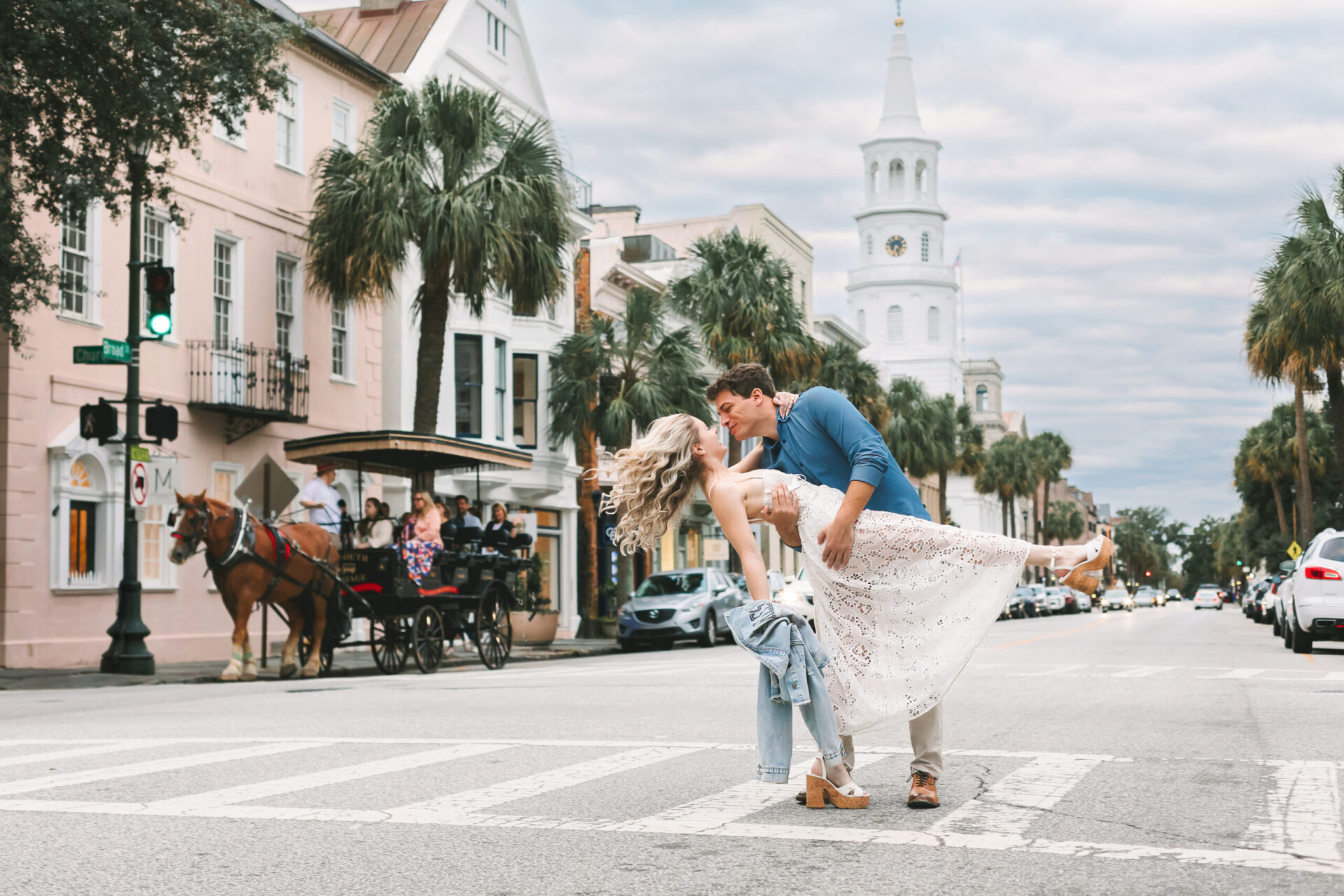 Elopement Wedding A couple is dancing in the middle of a crosswalk on a city street. The man, in a blue shirt and beige pants, is dipping the woman, who wears a white dress. A horse-drawn carriage, parked cars, pedestrians, and historic buildings, including a white clock tower, are in the background. Elopements Inc