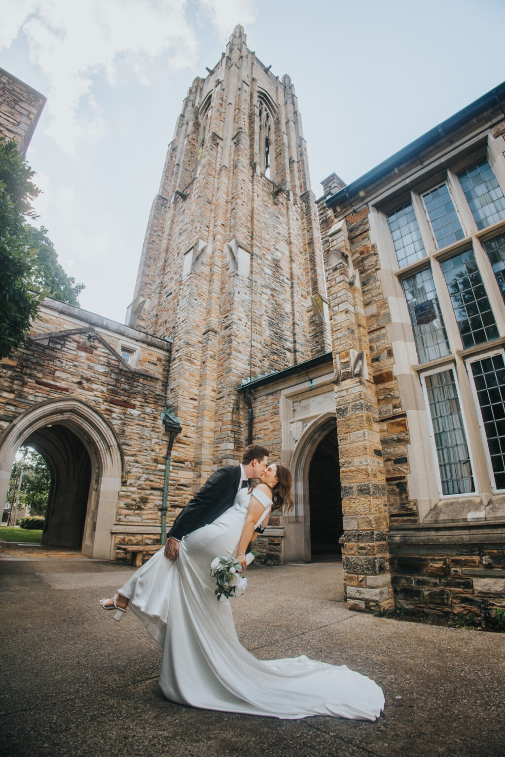 Elopement Wedding A bride and groom, eloping in a romantic escape, share a kiss in front of a large stone church with tall arched windows and a towering steeple. The groom, in a black suit, dips the bride, who is in a white gown and holding a bouquet. Set outdoors under a partly cloudy sky, both look joyous. Elopements Inc