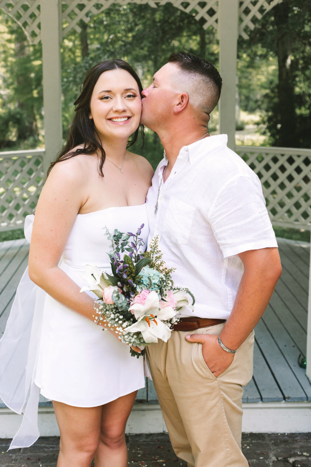 Elopement Wedding A couple stands in front of a gazebo. The woman, in a white strapless dress, holds a bouquet of flowers and smiles at the camera. The man, wearing a white shirt and khaki pants, stands beside her, affectionately kissing her on the cheek. Their elopement is set against a lush backdrop of greenery. Elopements Inc