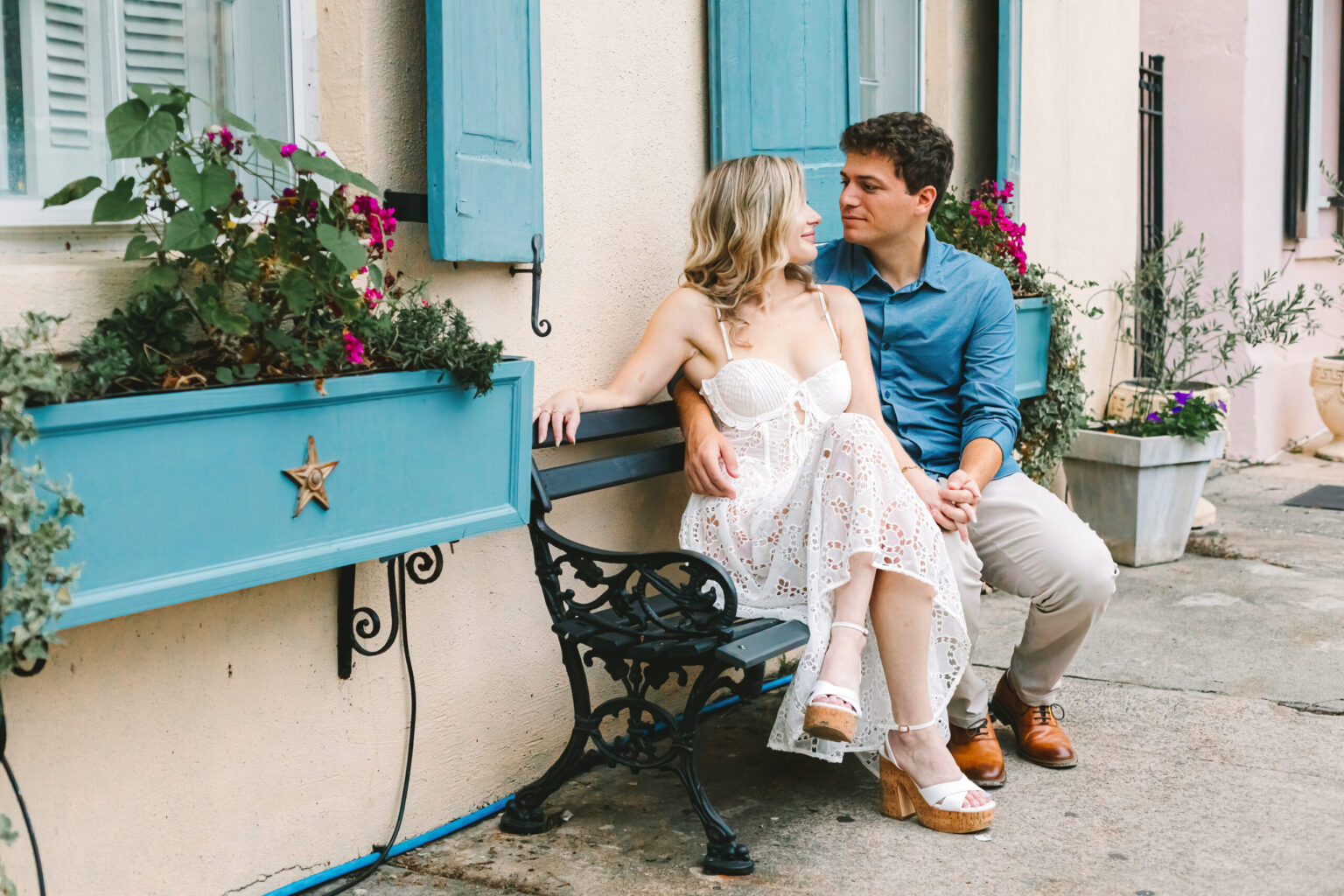 Elopement Wedding A couple sits on a black wrought-iron bench in front of a beige house with blue shutters and a flower box. The woman wears a white dress and platform sandals, while the man is in a blue long-sleeve shirt and khaki pants. They look at each other affectionately, as if savoring the joy of their recent elopement. Potted plants decorate the area. Elopements Inc