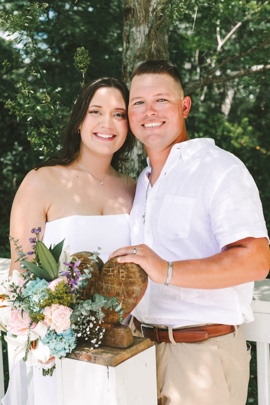 Elopement Wedding A smiling couple stands closely together outdoors, dressed in light-colored clothing. The woman, wearing a white dress, holds a bouquet of flowers. The man, in a white shirt and khaki pants, rests his hand on a carved wooden heart on a pedestal. Greenery and trees are visible in the background—a perfect setting for an intimate elopement. Elopements Inc