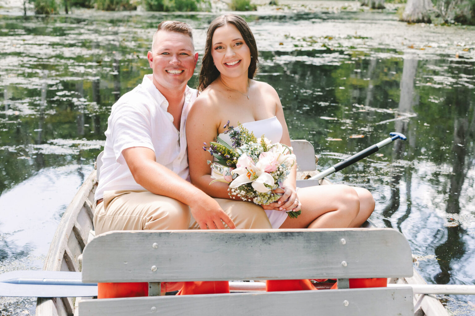 Elopement Wedding A smiling couple sits in a small rowboat on a serene lake, surrounded by lush greenery. The man, in a white shirt and beige pants, has his arm around the woman, who is wearing a white dress and holding a bouquet of flowers. The tranquil water reflects their intimate elopement in this peaceful outdoor setting. Elopements Inc