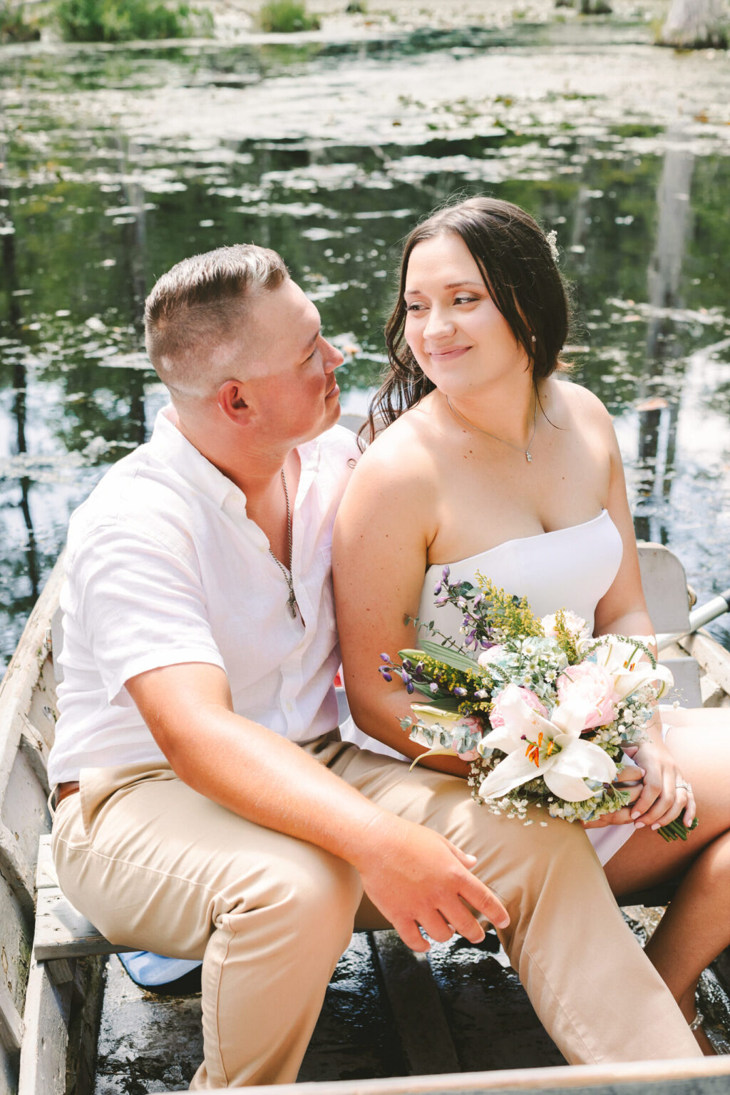 Elopement Wedding A couple sits closely in a small boat on a calm lake, their elopement unfolding in serene beauty. The man, wearing a white shirt and khaki pants, gazes at the woman in her strapless white dress holding a colorful bouquet. Green foliage and lily pads create a picturesque backdrop for their intimate moment. Elopements Inc