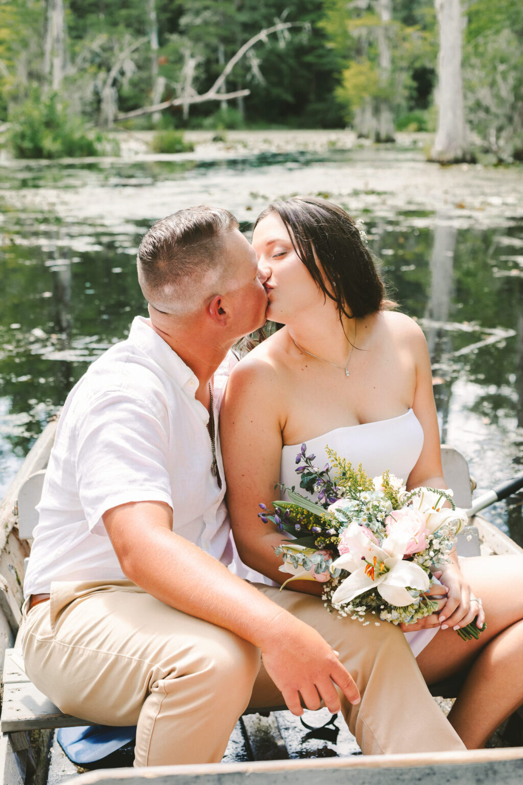 Elopement Wedding A couple eloping sits in a rustic rowboat on a serene lake surrounded by greenery. They are sharing a kiss, and the woman holds a bouquet of flowers featuring white lilies. The man wears a light-colored short-sleeve shirt and khaki pants, while the woman is in a white strapless dress. Elopements Inc