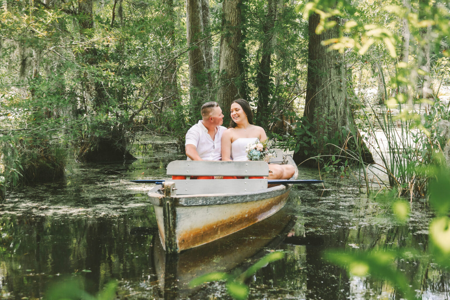 Elopement Wedding A couple elopes in a small boat in a serene, wooded swamp. The man wears a white shirt and jeans, while the woman, in a white strapless dress, holds a bouquet. They are smiling and looking at each other. The boat is surrounded by lush greenery and still water, creating a peaceful, intimate setting. Elopements Inc