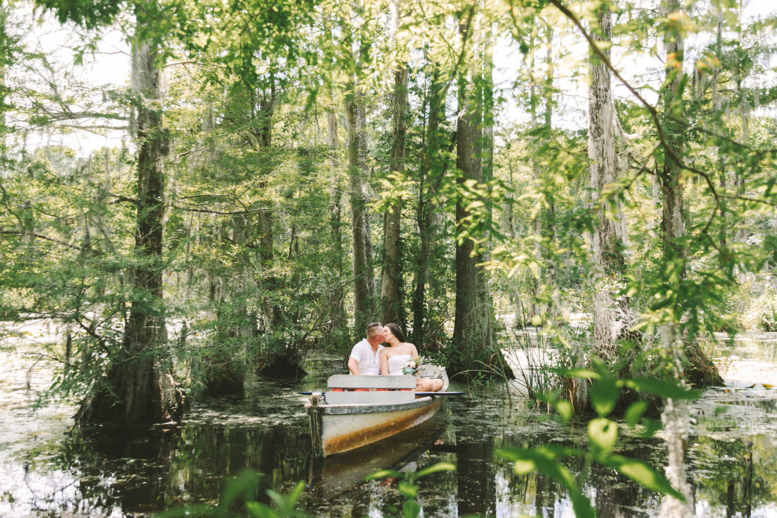 Elopement Wedding A couple sits in a canoe embraced in each other's arms, sharing a kiss in a swampy, lush forest. Sunlight filters through the dense foliage of tall, moss-covered trees that surround them, creating a serene, secluded atmosphere. The water reflects the greenery, an idyllic spot for elopements. Elopements Inc