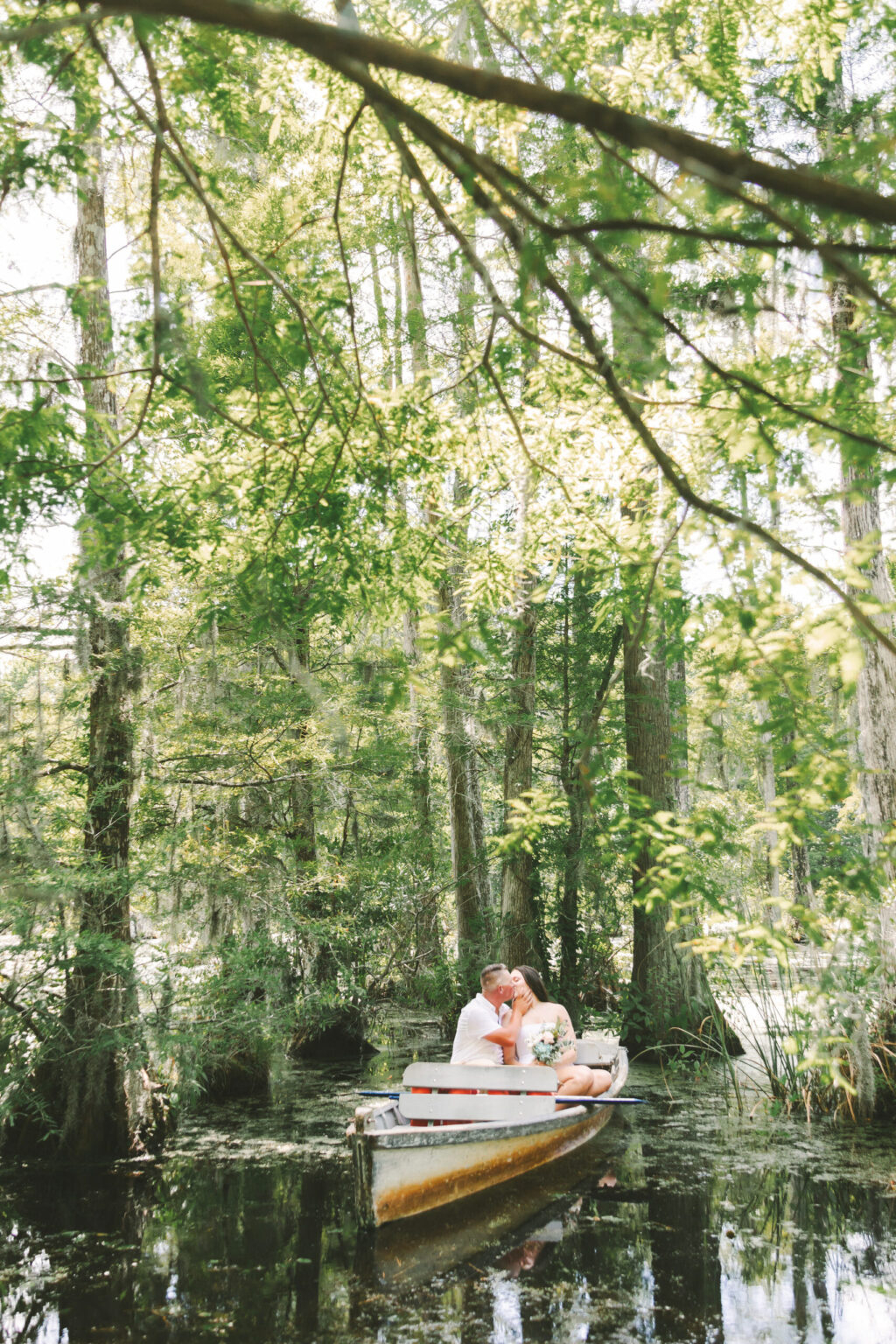 Elopement Wedding A couple sits in a small boat, sharing an embrace and kiss amidst a lush, green forest. They are surrounded by tall trees with thick foliage, reflecting on the calm water below. Sunlight filters through the leaves, casting dappled light on the serene elopement scene. Elopements Inc