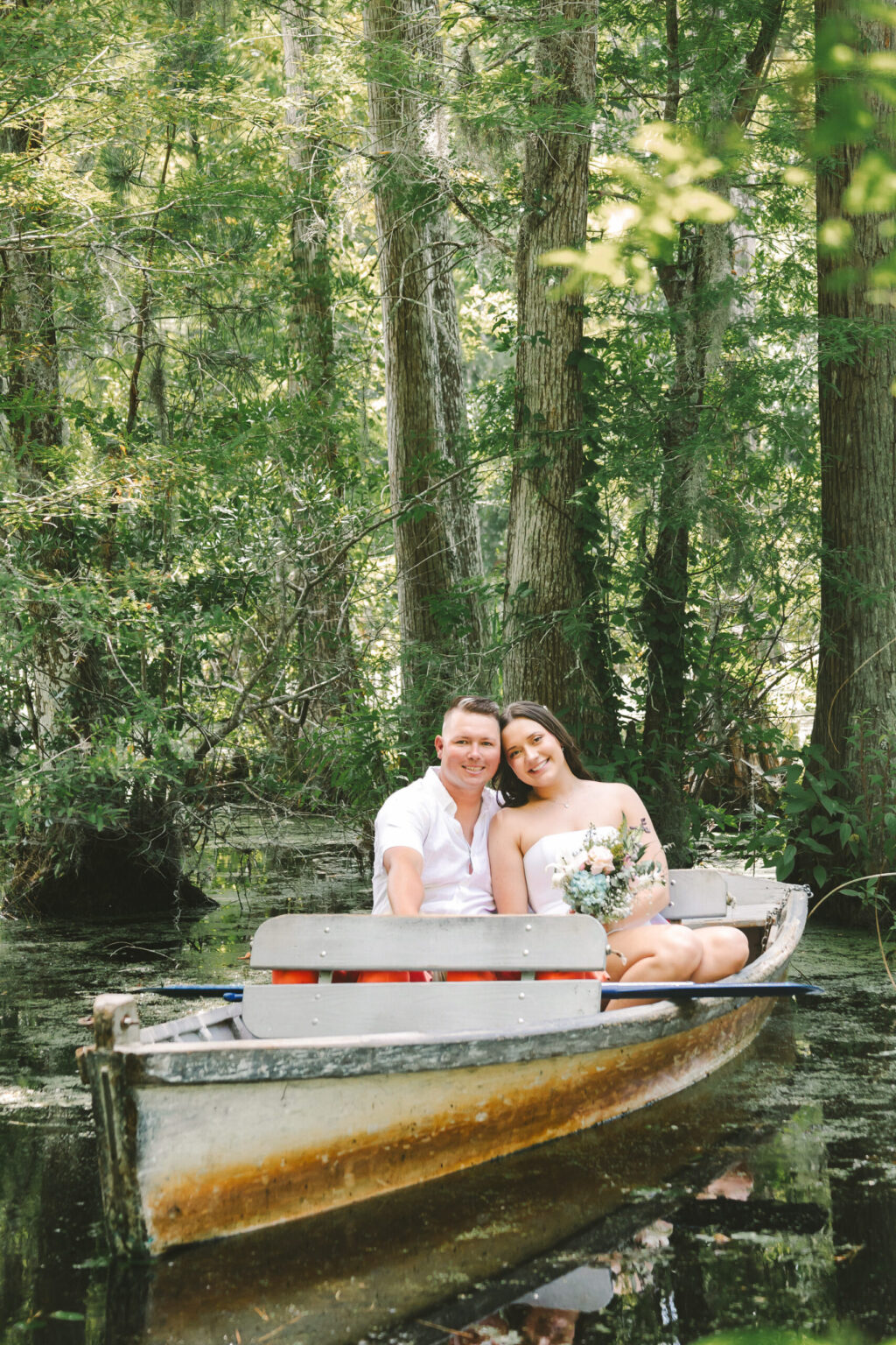 Elopement Wedding A couple, eloping in a lush, green forest setting, sit in a small boat. The man is dressed in a white shirt and the woman in a white dress, holding a bouquet. They smile at the camera as their boat floats on calm water surrounded by trees. Elopements Inc