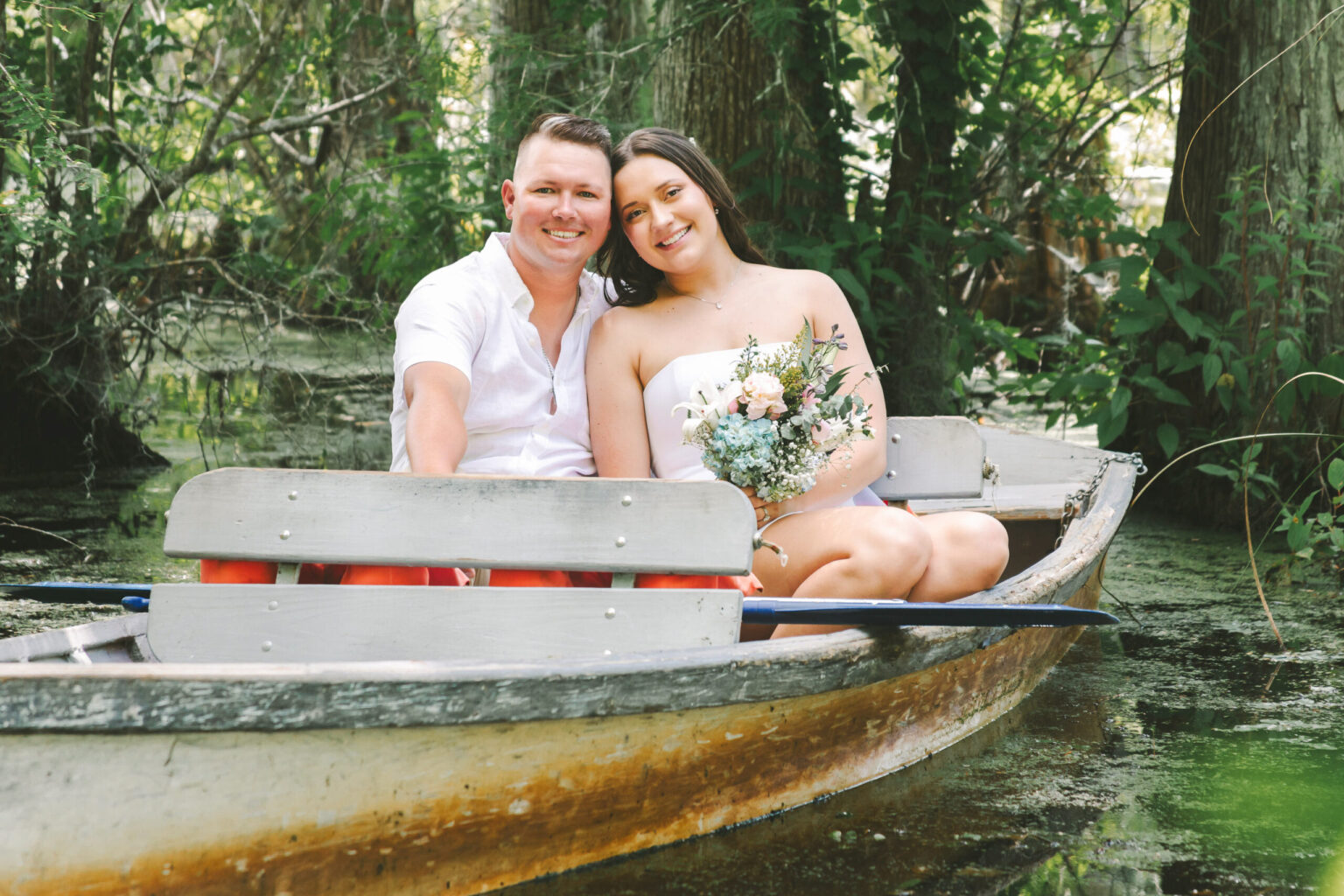 Elopement Wedding A couple sits in a small wooden boat on a narrow, calm, tree-lined waterway. Both are dressed casually; the man in a white shirt and the woman in a white dress holding a bouquet of flowers. They smile warmly at the camera, eloping amidst the lush greenery on a partly cloudy day. Elopements Inc