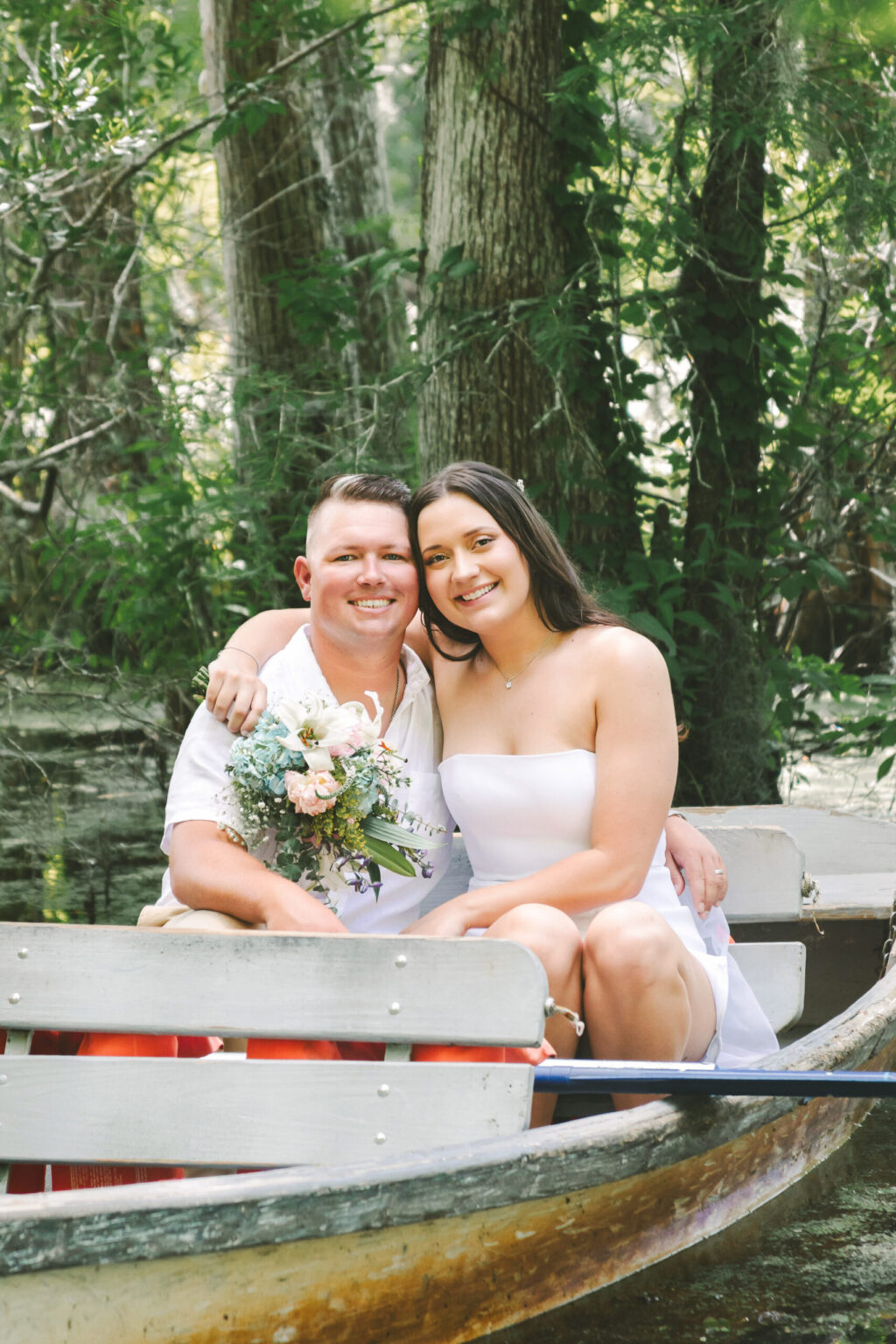 Elopement Wedding A couple sits in a canoe on a calm, wooded river, eloping amidst nature's beauty. The man, wearing a white shirt, holds a bouquet of flowers and has his arm around the woman. She is dressed in a strapless white top and has long brown hair. Both are smiling and surrounded by lush greenery, creating a serene, romantic atmosphere. Elopements Inc