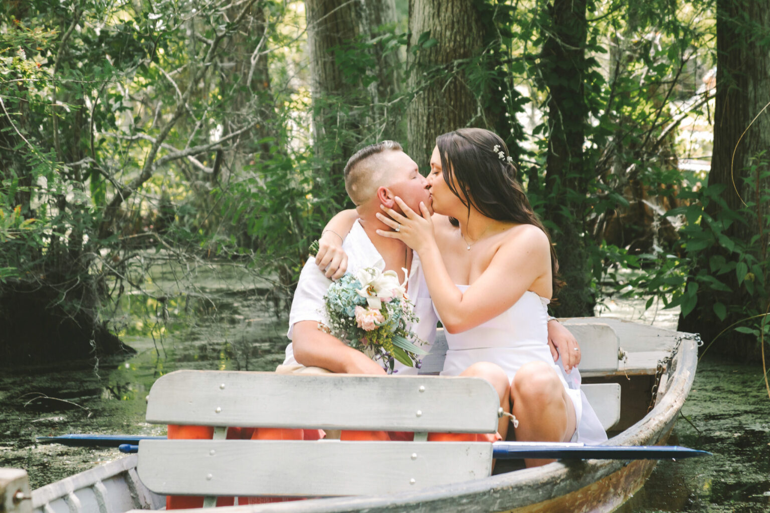 Elopement Wedding A couple sits in a wooden boat on a forested lake. The woman, wearing a white dress, lovingly kisses the man holding a bouquet of flowers. They are surrounded by lush green trees and vegetation, creating an intimate setting perfect for romantic elopements. Elopements Inc