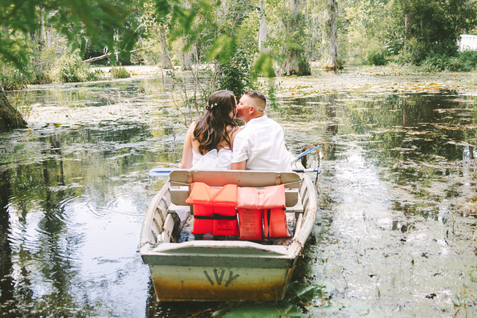 Elopement Wedding A couple sits in a small rowboat, embraced and kissing, on a serene lake surrounded by lush greenery and trees. The boat has two bright red life jackets attached at the back. Eloping amidst nature's beauty, the water is calm, covered with patches of lily pads and aquatic plants. The couple appears relaxed and in love. Elopements Inc