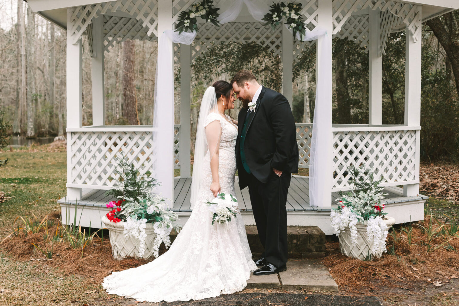 Elopement Wedding A bride and groom stand facing each other under a white gazebo. The bride, in a lace gown with a long train and veil, holds a bouquet of white flowers. The groom, in a black suit, has his forehead touching the bride's. Floral arrangements in white and red decorate the gazebo steps—perfect for an intimate elopement. Elopements Inc