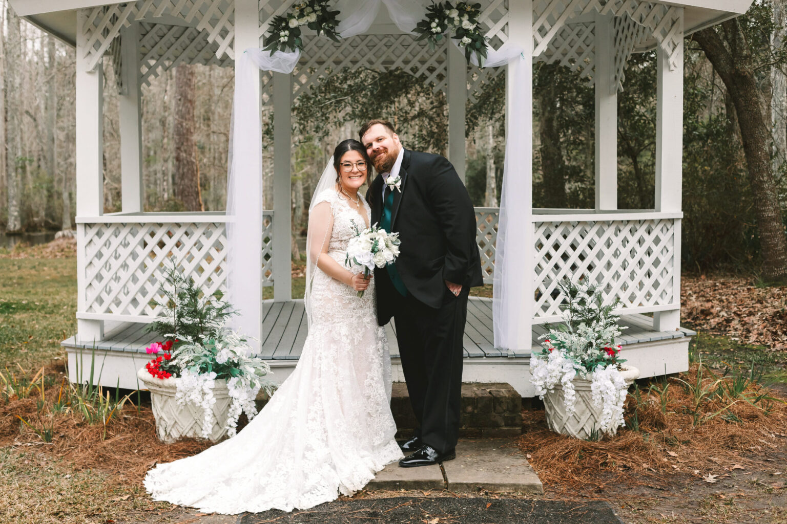 Elopement Wedding A bride in a lacy white gown holding a bouquet stands next to a groom in a black suit and tie. They are smiling and standing close together in front of a white gazebo adorned with white drapery and greenery, the perfect setting for their intimate elopement. The scene is set outdoors with trees in the background. Elopements Inc