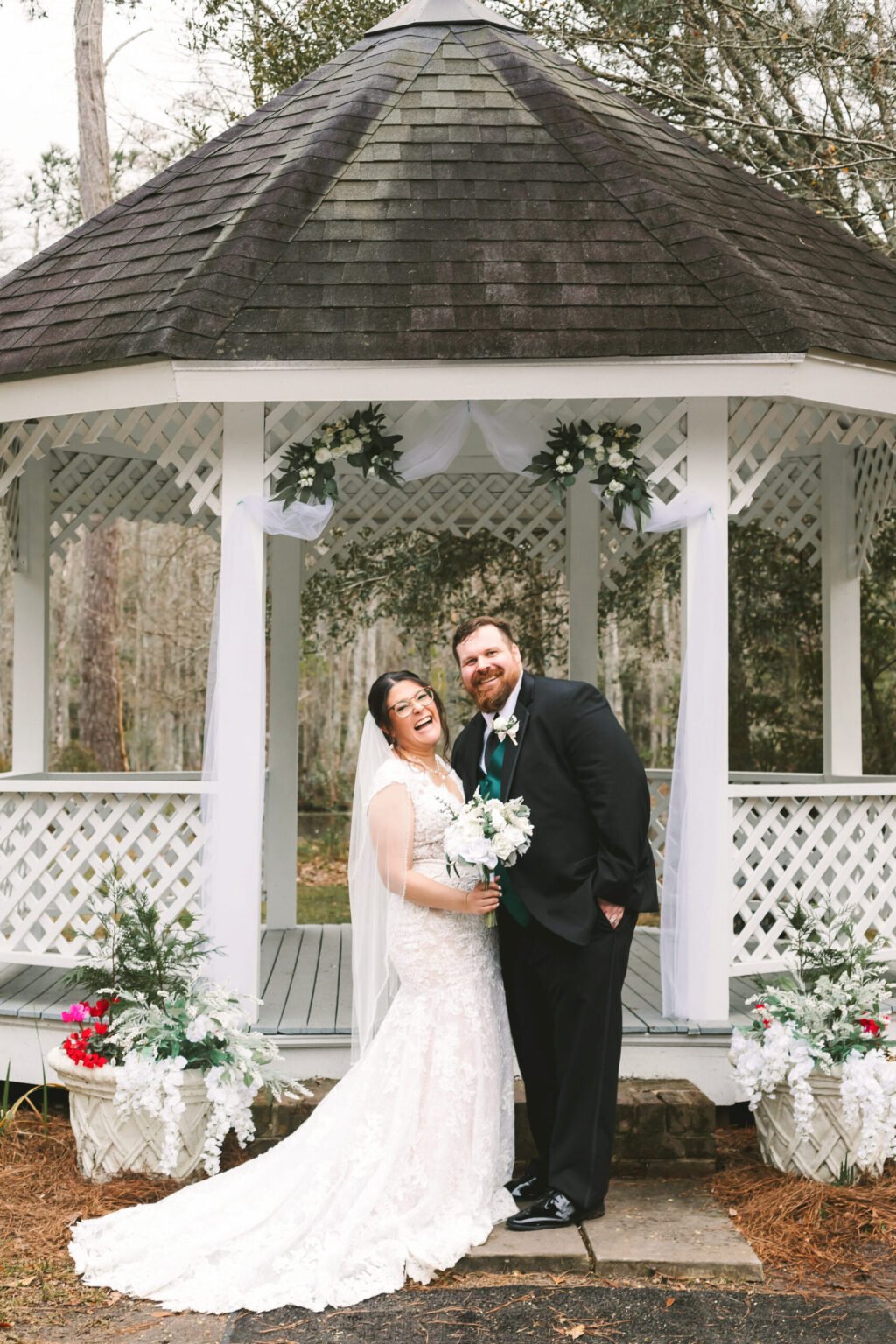 Elopement Wedding A bride and groom stand smiling under a white gazebo decorated with white flowers and sheer fabric, perfect for intimate elopements. The bride wears a lace gown and holds a white bouquet, while the groom is in a black suit with a green tie. Greenery and red flowers surround the gazebo, with trees visible in the background. Elopements Inc