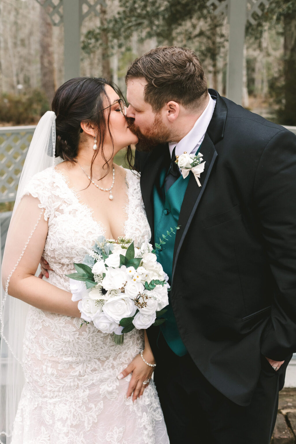 Elopement Wedding A bride and groom share a kiss. The bride, in a white lace wedding dress, holds a bouquet of white flowers. The groom wears a black suit with a teal vest and white boutonniere. This intimate elopement takes place outdoors near a decorative structure, with the bride donning both veil and glasses. Elopements Inc