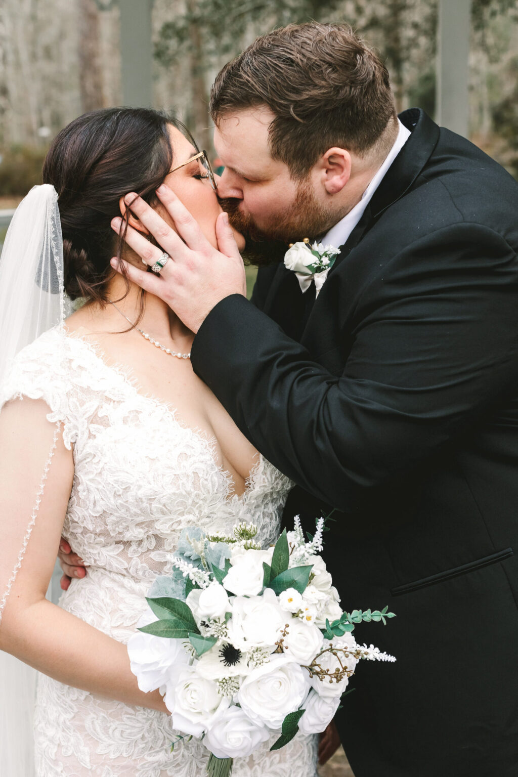 Elopement Wedding A newlywed couple shares a kiss on their elopement day. The bride, in a lace dress and veil, holds a bouquet of white flowers. The groom, wearing a black suit and a boutonniere, lovingly holds the bride's face with his hand. They stand outdoors with a blurry natural background. Elopements Inc