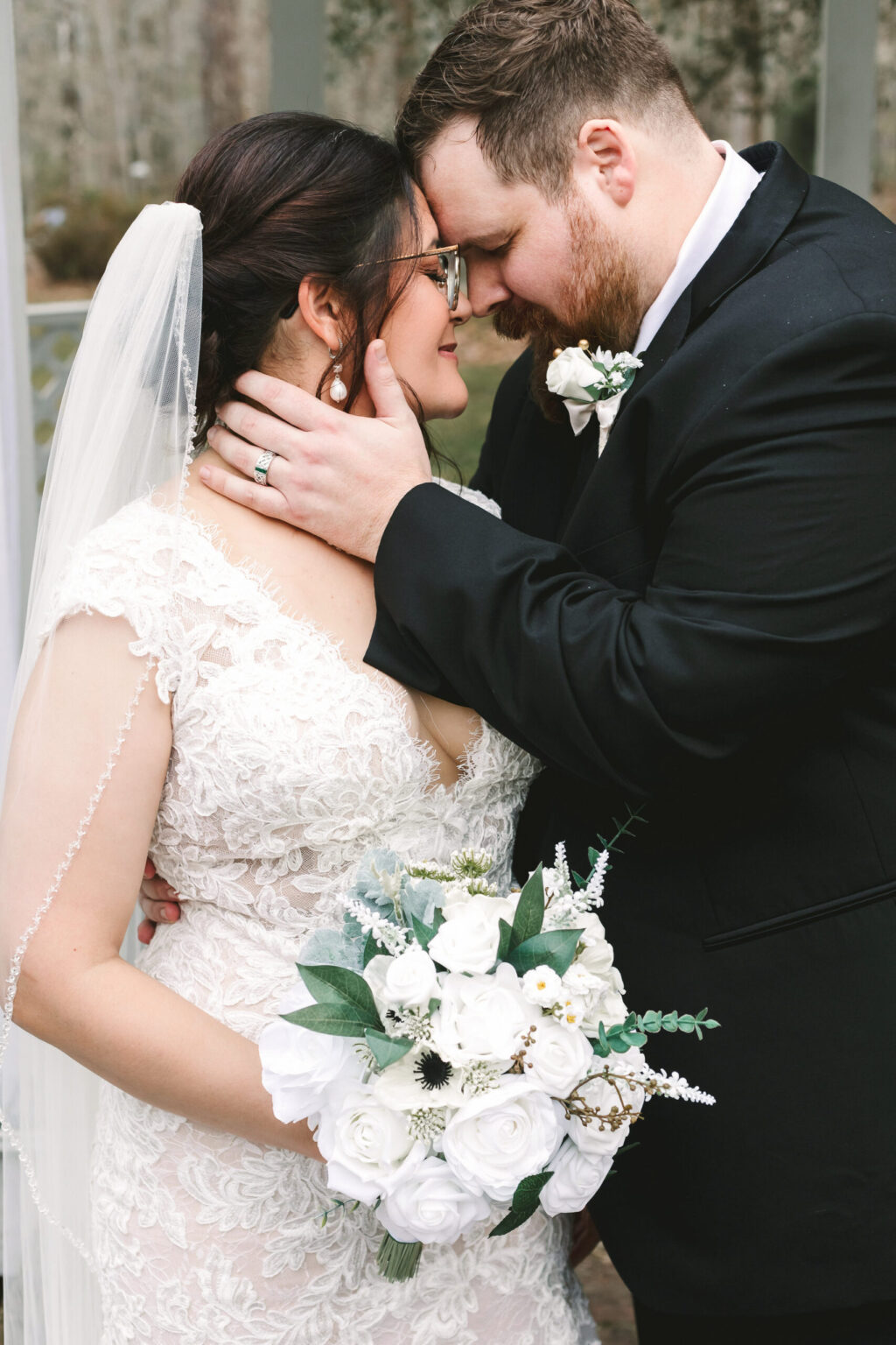 Elopement Wedding A bride in a white lace wedding dress and veil holds a bouquet of white roses, greenery, and small flowers. The groom, in a black suit with a white shirt and boutonniere, tenderly cups her face and leans his forehead against hers. Their closed eyes and affectionate expressions capture the intimacy of their elopement. Elopements Inc