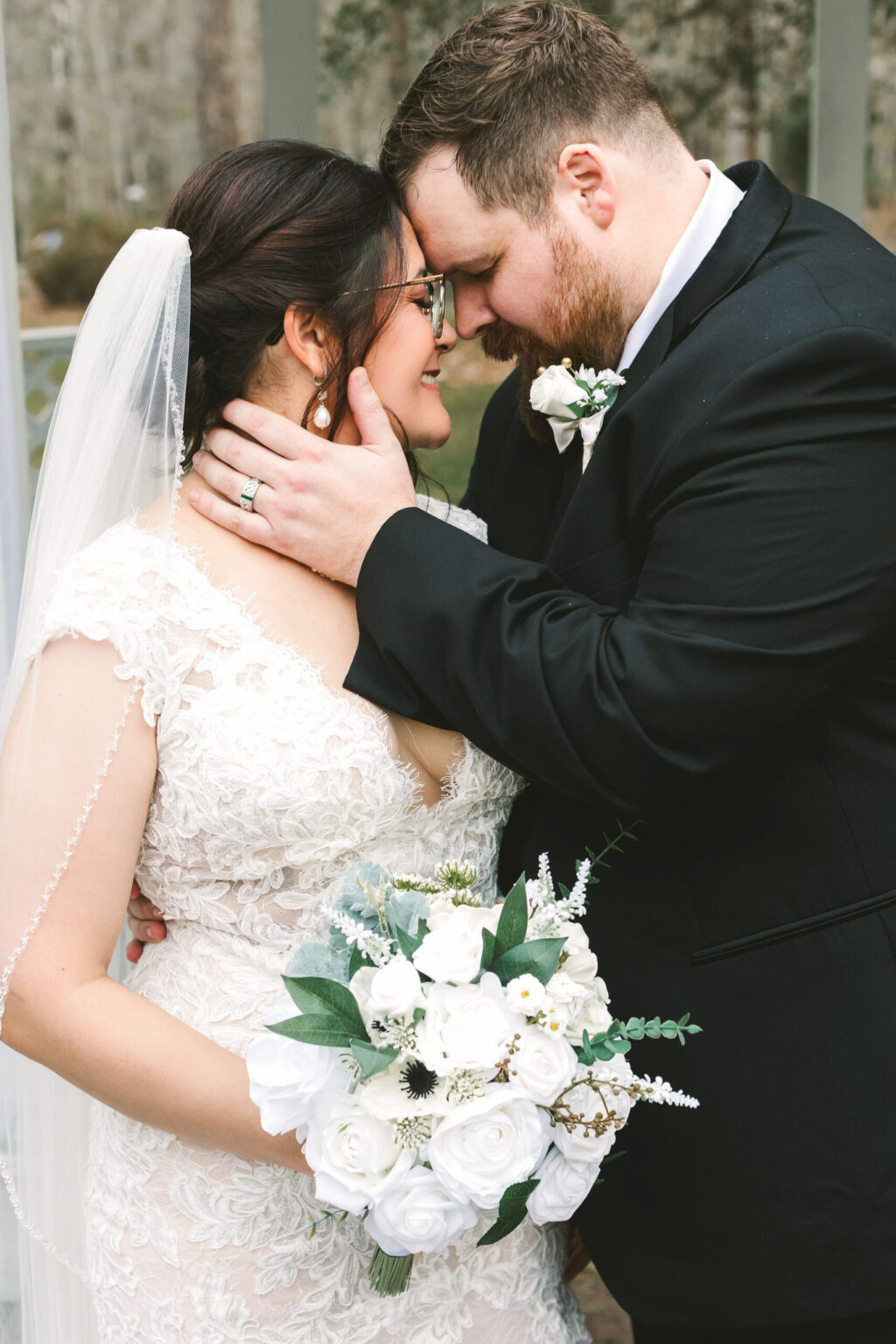 Elopement Wedding A bride and groom share an intimate moment, touching foreheads and smiling. The bride has dark hair, wears glasses, a lace gown, and holds a bouquet of white flowers. The groom has light hair, wears a black suit with a white boutonniere, and gently holds the bride's face with one hand—capturing the essence of their elopement. Elopements Inc