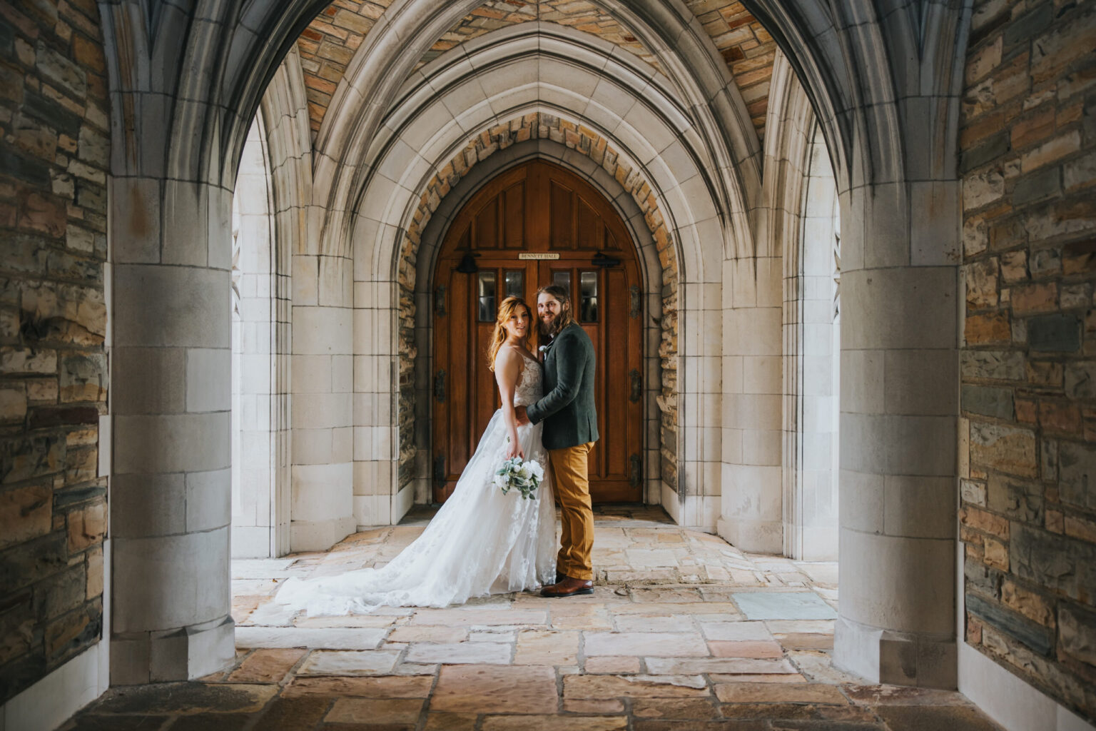 Elopement Wedding A bride in a white gown and a groom in a green jacket and brown trousers stand under a stone archway with gothic architectural elements. The bride holds a bouquet, and they both look towards the camera, standing on a stone floor, with wooden double doors visible in the background—a perfect scene for an intimate elopement. Elopements Inc