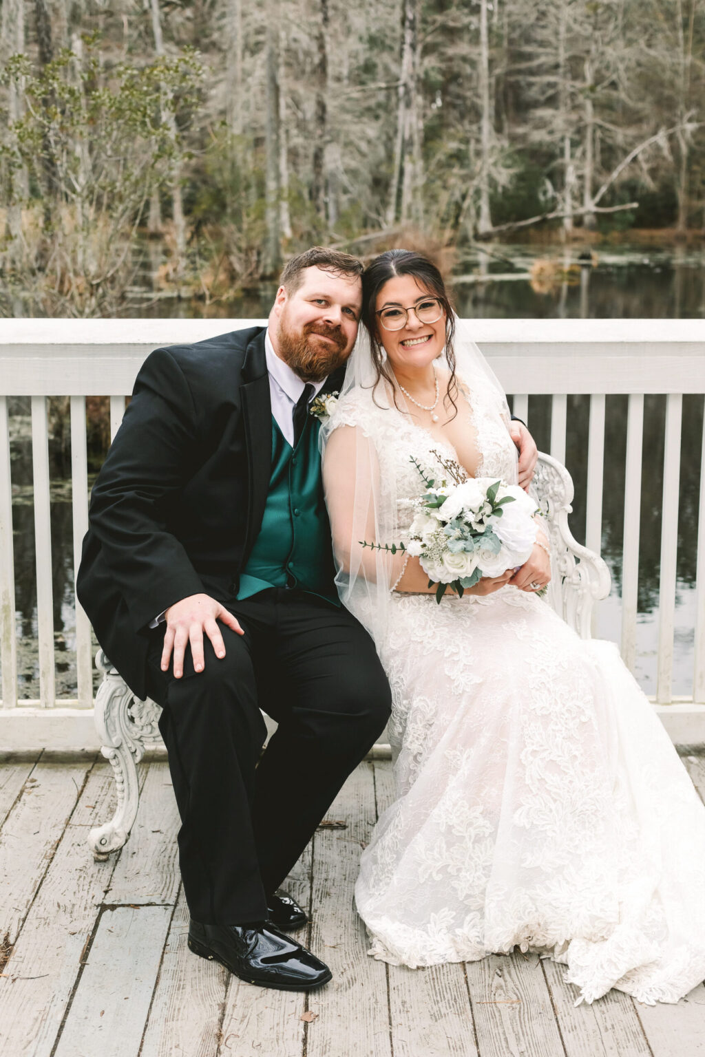 Elopement Wedding A bride in a lace wedding gown and veil sits on a white bench next to a groom in a black suit with a green vest and tie. Both are smiling and posing for the photo. Behind them is a wooden deck with a white railing, overlooking a serene pond and forested area—perfect for an intimate elopement. Elopements Inc