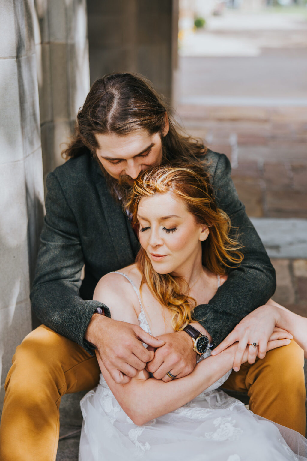 Elopement Wedding A man with long, brown hair embraces a woman with long, reddish hair from behind. Seated on a stone step, the woman in a white dress and the man in a dark jacket and tan pants—they share a tender moment, contemplating their imminent elopement with serene expressions. Elopements Inc