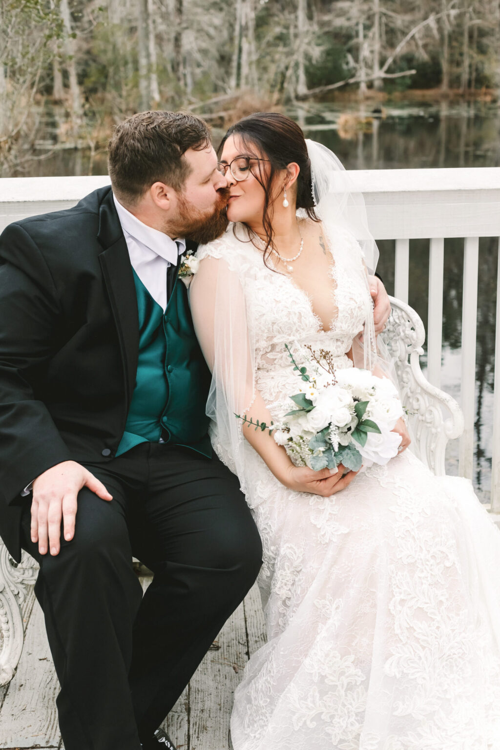 Elopement Wedding A bride and groom share a kiss while sitting on a white bench outside near a lake, capturing the magic of their elopement. The bride, in a white lace dress and veil, holds a bouquet of white flowers. The groom, in a black suit with a teal vest and tie, has his arm around her. Trees and water create a serene background. Elopements Inc