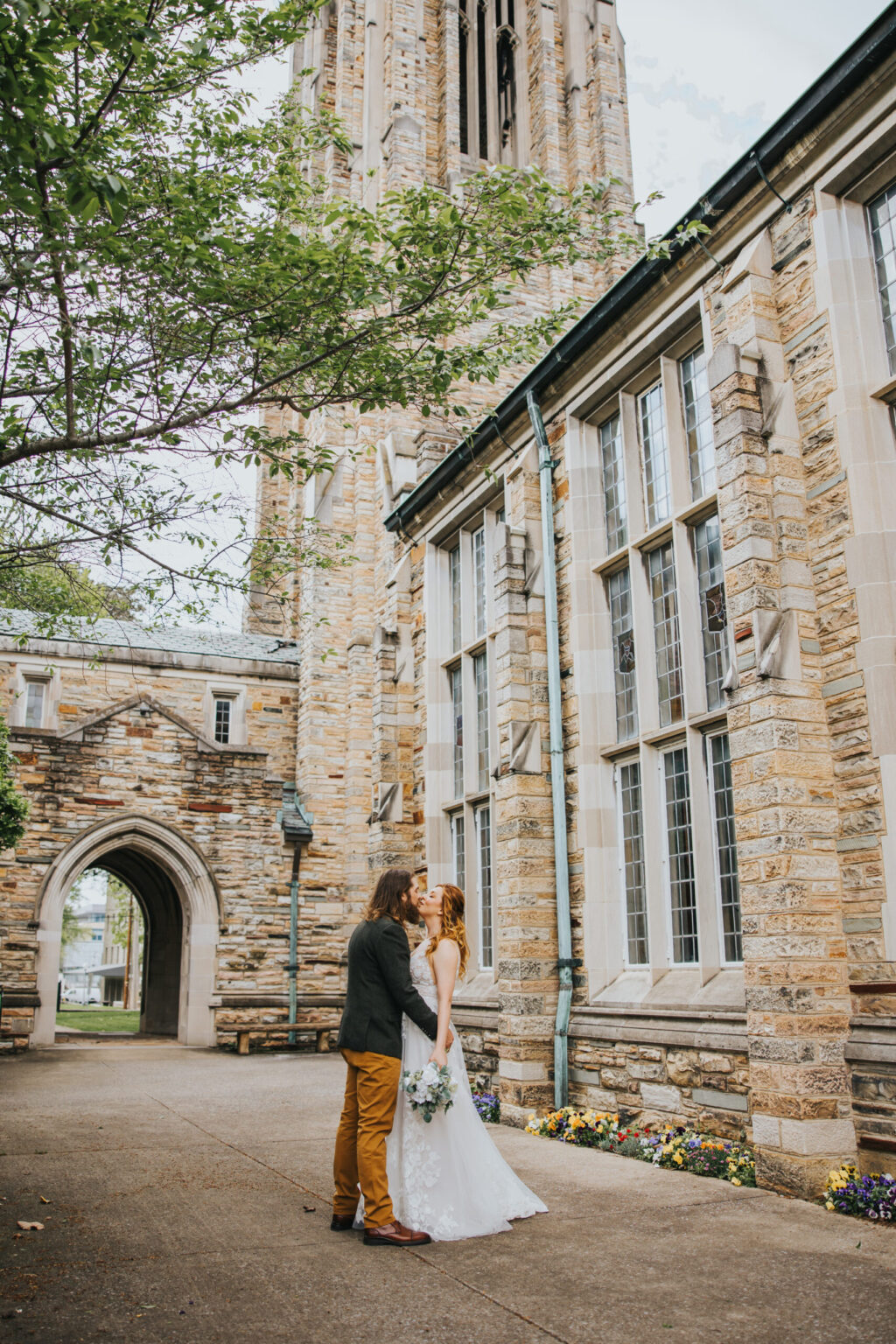 Elopement Wedding A couple stands outside a stone building with tall windows and an arched entrance. The man, in a grey blazer and brown pants, kisses the woman, who is in a white wedding dress and holding a bouquet. Framed by greenery and a tall tower in the background, this intimate elopement captures pure romance. Elopements Inc