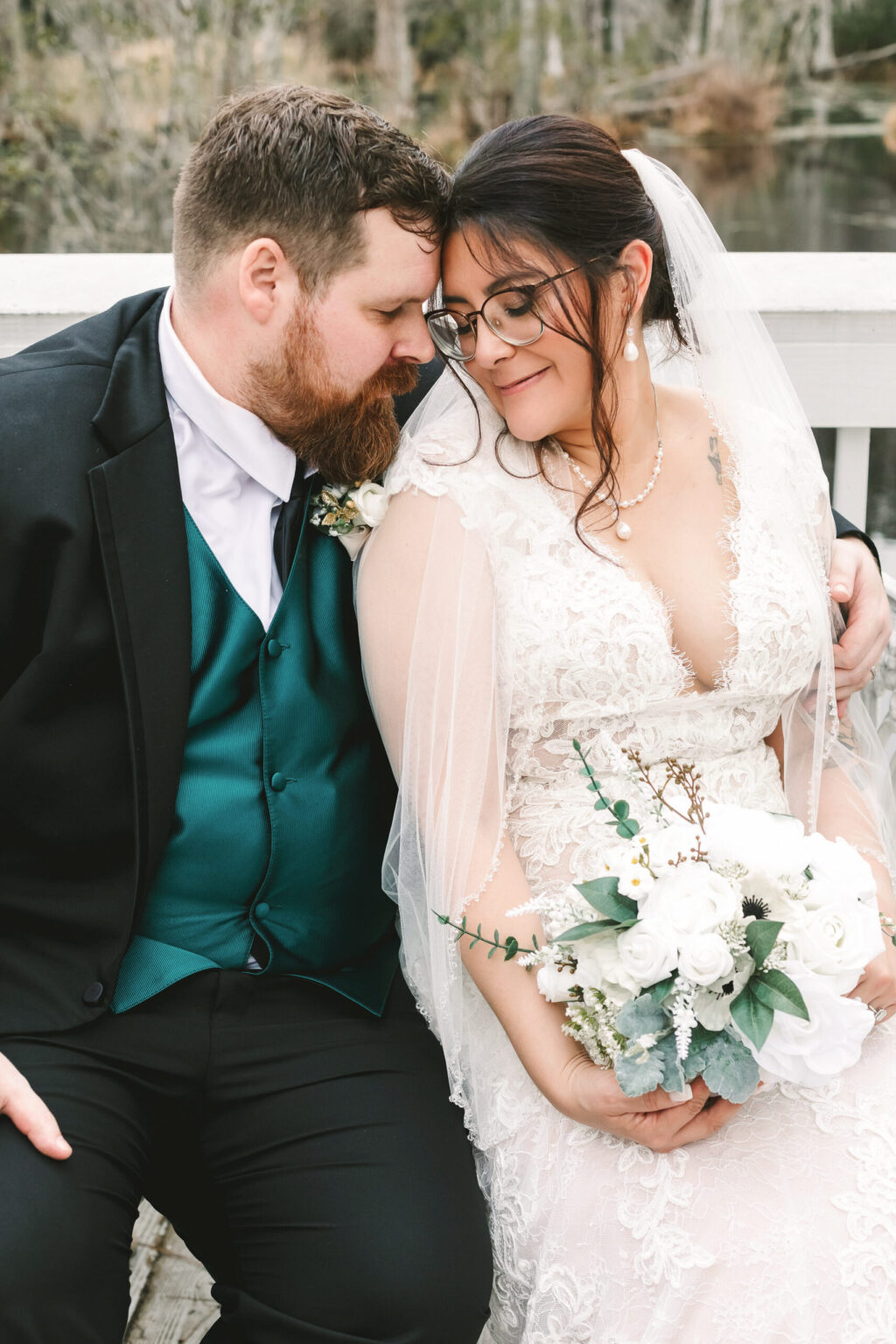 Elopement Wedding A bride and groom sit closely on a bench outside near a body of water, enjoying their elopement. The groom, with a beard, wears a black suit with a teal vest and tie. The bride, in a lace gown and veil, holds a bouquet of white flowers and greenery. They touch heads gently, eyes closed, sharing a tender moment. Elopements Inc