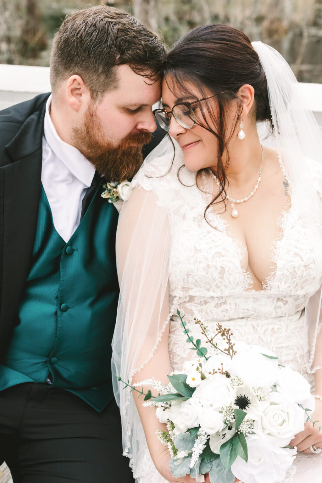 Elopement Wedding A bride and groom share an intimate moment during their elopement. The groom, in a dark suit, white shirt, and teal vest, rests his head gently against the bride's. The bride, in a lace wedding dress with a veil and glasses, holds a white floral bouquet and wears a pearl necklace and earrings. Both look content and happy. Elopements Inc