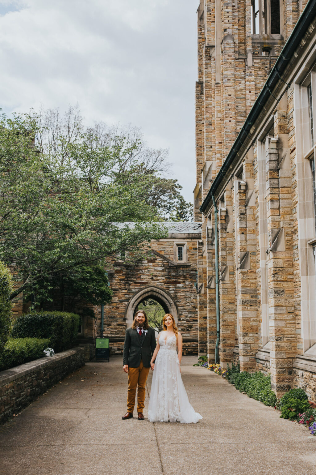 Elopement Wedding A couple stands outdoors in front of a large stone building with arched windows and greenery. The man wears a dark jacket and tan pants while the woman is in a white lace wedding dress. They hold hands and smile at the camera, capturing the joy of their intimate elopement. Elopements Inc