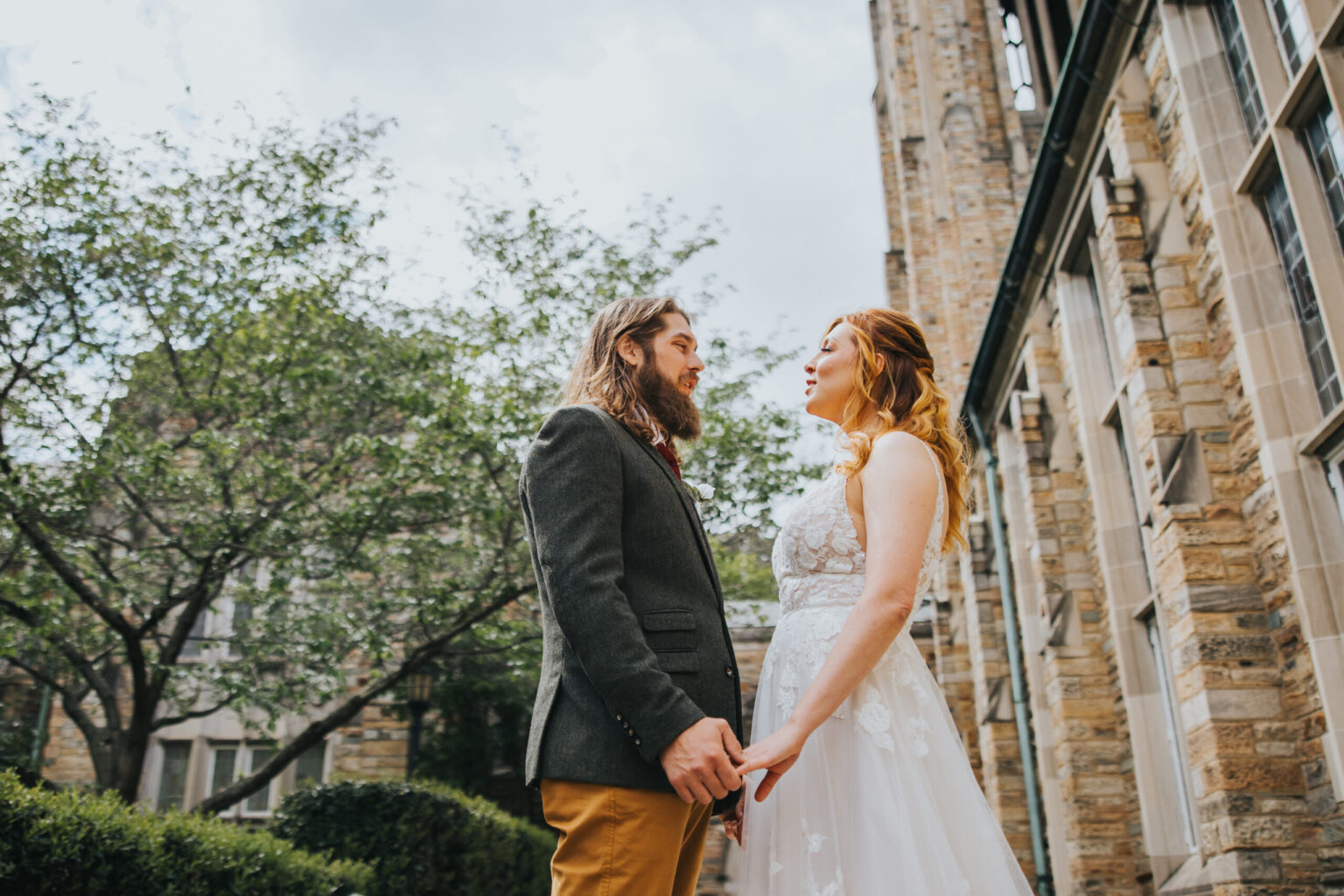 Elopement Wedding A couple holding hands stands outdoors near a stone building with arched windows, surrounded by greenery. The man has long hair, a beard, and wears a dark jacket with mustard trousers. The woman has red hair and wears a white, sleeveless dress. Both are gazing at each other happily, as if ready to elope. Elopements Inc