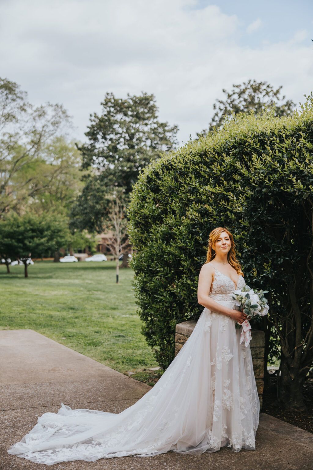 Elopement Wedding A bride with long strawberry blonde hair stands beside a lush green hedge, wearing a strapless, white lace wedding gown with a long train. She holds a bouquet of white and green flowers. The scene is outdoors on a cloudy day with trees and grass in the background, perfect for intimate elopements. Elopements Inc