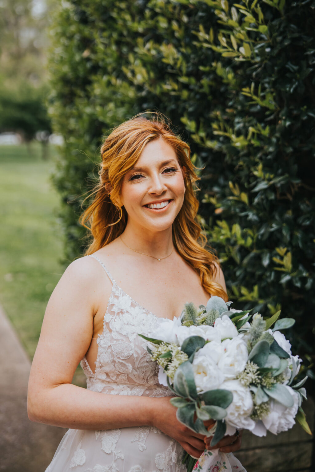 Elopement Wedding A woman in a white, floral lace wedding dress poses outdoors with a bouquet of white flowers and green foliage. She has light brown, wavy hair and is smiling. Behind her is a tall, green hedge. The background shows a grassy area and trees, capturing the intimate beauty of an elopement setting. Elopements Inc