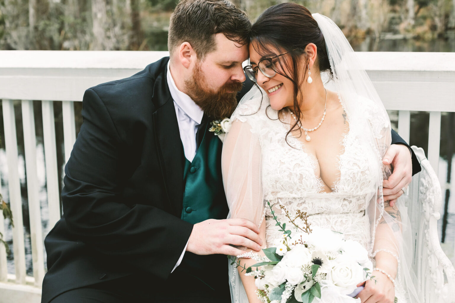 Elopement Wedding A bride and groom sit closely together on a white bench, smiling and leaning towards each other. The bride, likely having chosen to elope, wears a white lace dress with a veil and holds a bouquet of white flowers. The groom is in a black suit with a dark green vest. They look content and happy. A wooden railing and trees are in the background. Elopements Inc