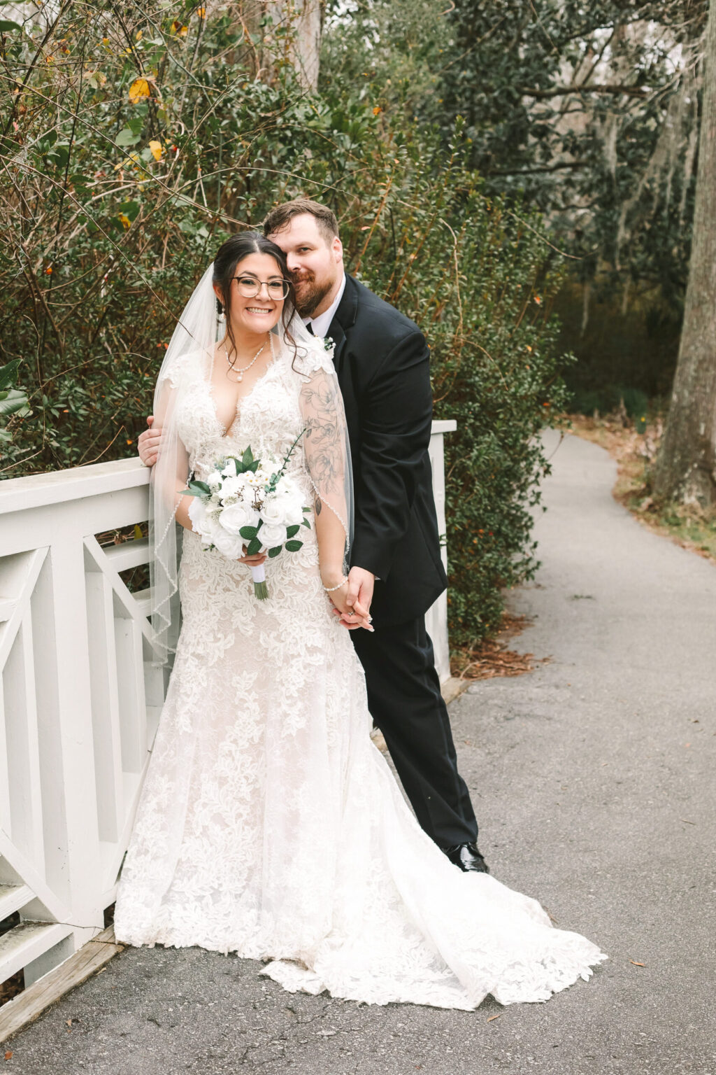 Elopement Wedding A couple in wedding attire stands on a paved path beside a white wooden fence, surrounded by greenery. The bride, in a lace gown and veil, holds a bouquet of white flowers. The groom, in a black suit, affectionately stands behind her as they smile at the camera—capturing their intimate elopement. Elopements Inc