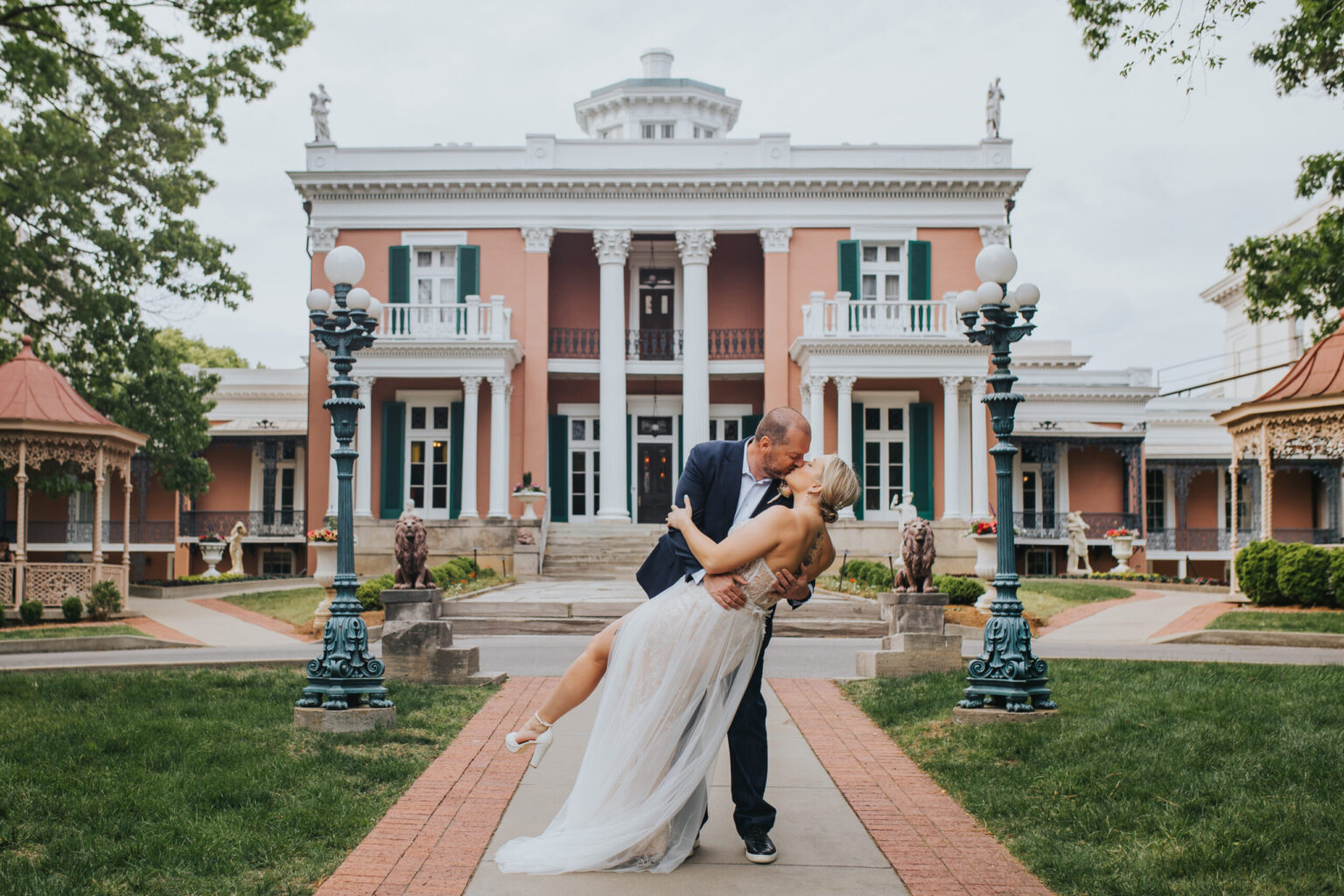 Elopement Wedding A newly married couple shares a kiss as the groom lifts the bride in front of a grand, historic mansion with white columns and multiple balconies. The groom is in a navy suit, and the bride is in a flowing white gown. They are surrounded by lush greenery and symmetrical walkways—perfect for an elopement. Elopements Inc