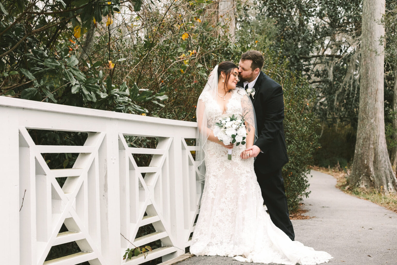 Elopement Wedding A bride and groom, eloping, stand on a white bridge outdoors, surrounded by greenery. The bride, in a lacy white gown and veil, holds a bouquet of white flowers. The groom, dressed in a black suit, leans in to kiss her cheek. Both are smiling, creating a romantic and intimate moment. Elopements Inc