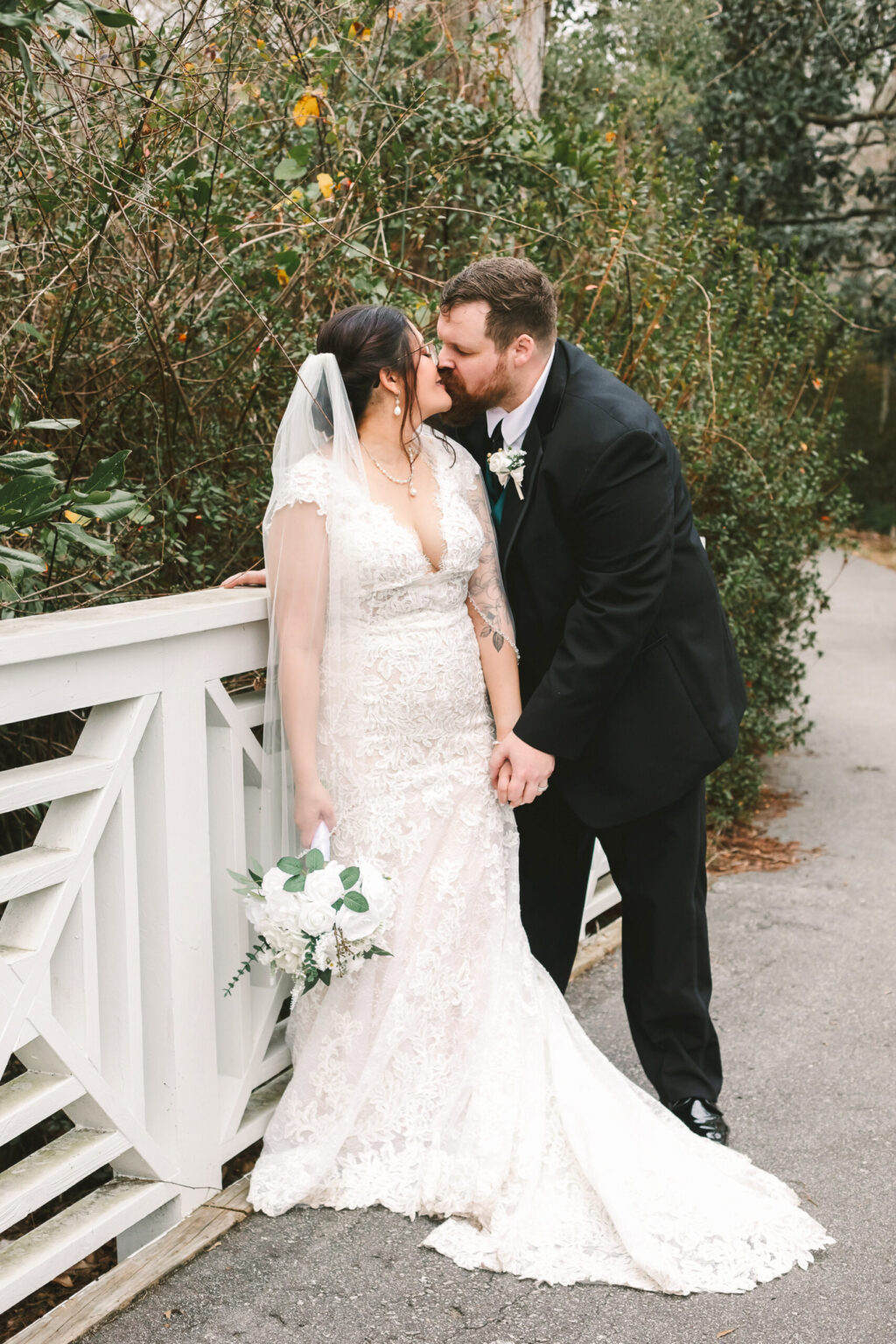 Elopement Wedding A couple, having eloped, stands close together on a pathway near greenery. The bride wears a lace wedding gown and holds a white floral bouquet, while the groom in a black suit leans in for a kiss. Both are smiling, enjoying their moment. A white fence is visible on the left. Elopements Inc