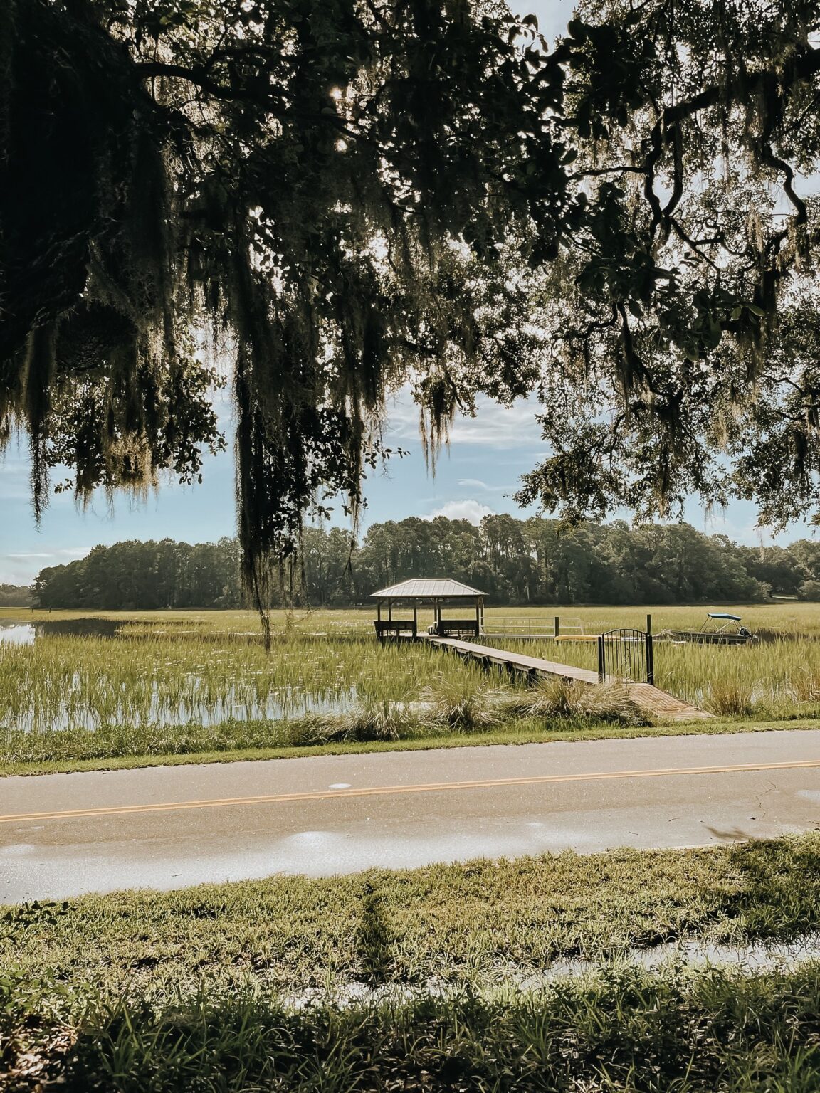 Elopement Wedding A tranquil scene of a wooden dock leading to a small pavilion over a marshy field with tall grass—an ideal spot for an intimate elopement. A road runs horizontally in the foreground, and hanging Spanish moss from a large tree frames the upper part of the image. Trees and a calm sky complete the background. Elopements Inc