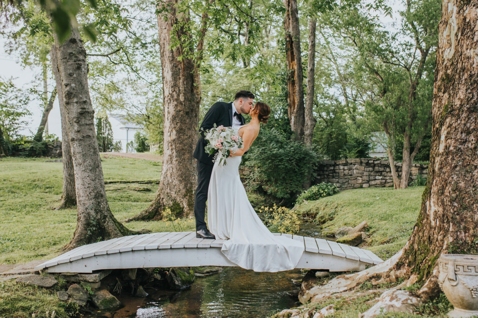 Elopement Wedding A bride and groom kiss on a small, white wooden bridge over a creek in a serene, green park. The bride wears a long, white gown and holds a bouquet, while the groom is dressed in a dark suit. Tall trees and lush greenery surround them, creating the perfect romantic setting for their elopement. Elopements Inc