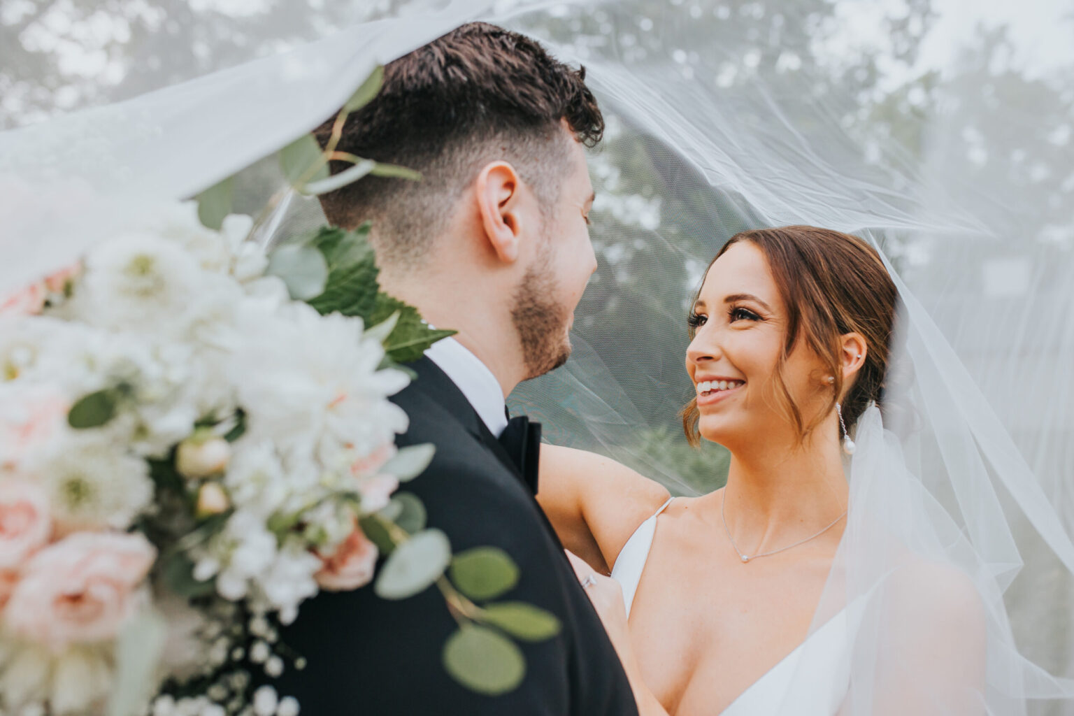 Elopement Wedding A bride and groom stand closely under a flowing white veil, looking at each other lovingly. The bride, with her hair up, holds a bouquet of white and pink flowers with green leaves. The groom is facing her, wearing a black suit and bow tie. The background is softly blurred with greenery—a perfect scene for elopements. Elopements Inc