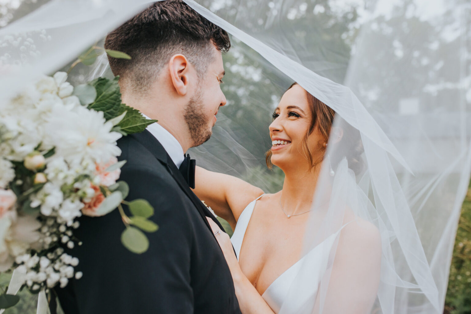 Elopement Wedding A bride and groom stand close under a veil, gazing into each other's eyes with joyful smiles. The bride is holding a white bouquet with green leaves and wears a white dress. The groom is in a black suit with a bow tie. The veil creates a soft, romantic atmosphere around their intimate elopement. Elopements Inc