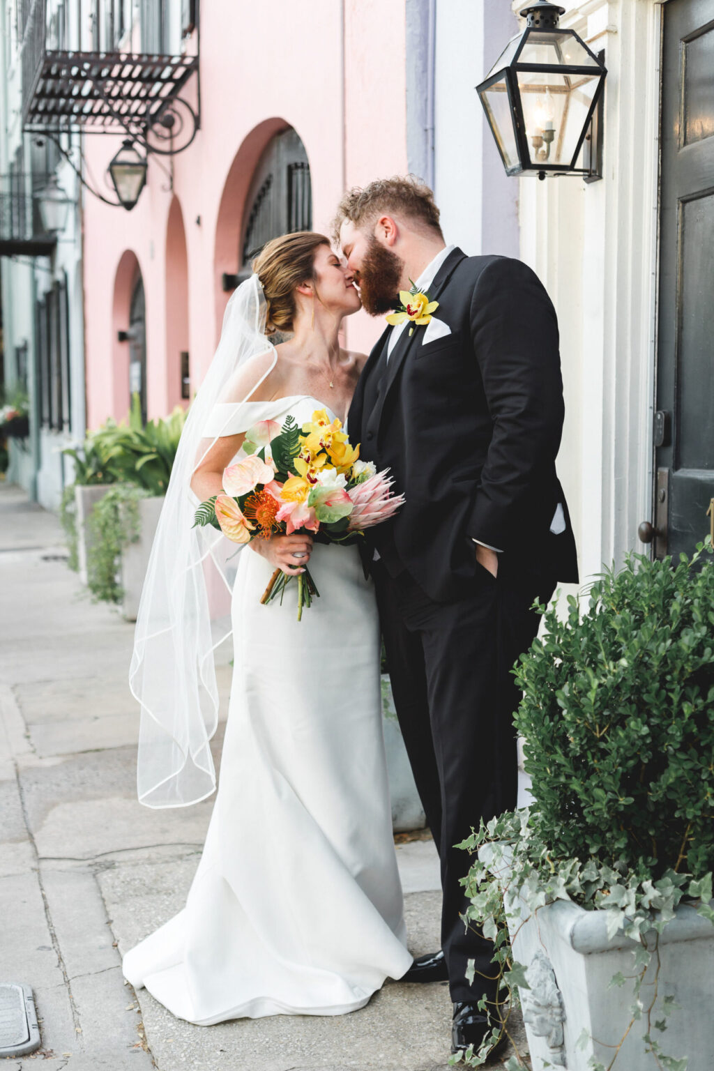 Elopement Wedding A bride in a white dress and veil holds a colorful bouquet while leaning in to kiss a groom in a black tuxedo. They stand on a street with pastel-colored buildings and black lanterns, perfect for intimate elopements. The groom's hand is in his pocket, and they are next to a door with potted plants nearby. Elopements Inc