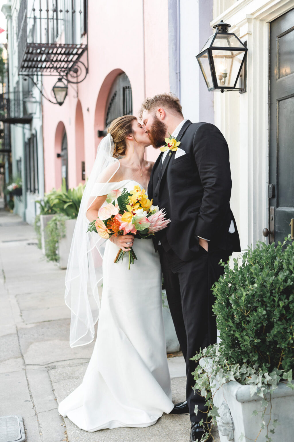 Elopement Wedding A bride in a white dress and veil holds a colorful bouquet, while a groom in a black suit and bow tie kisses her. They are eloping on a cobblestone street with pastel buildings and wrought-iron balconies in the background. The scene is romantic and picturesque. Elopements Inc