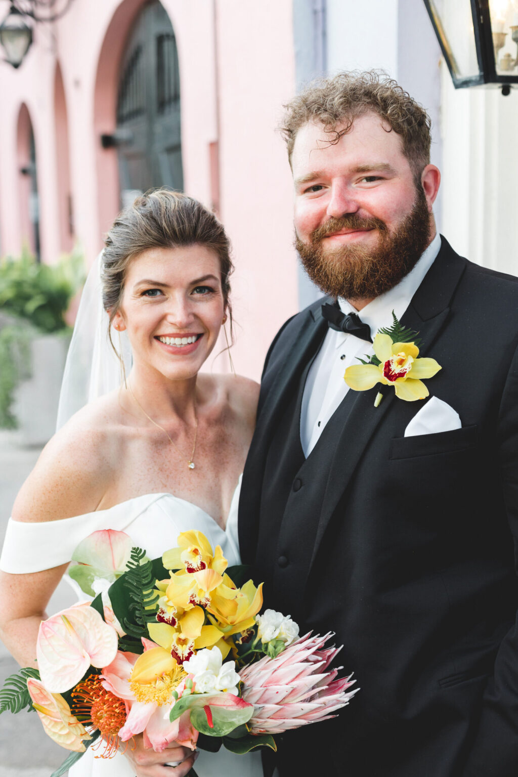 Elopement Wedding A smiling couple on their wedding day, possibly eloping, poses outdoors. The bride wears an off-the-shoulder white dress and veil, holding a colorful bouquet. The groom, with a beard, dons a black tuxedo and bowtie with a yellow flower boutonnière. A pastel pink building frames their joyous moment. Elopements Inc